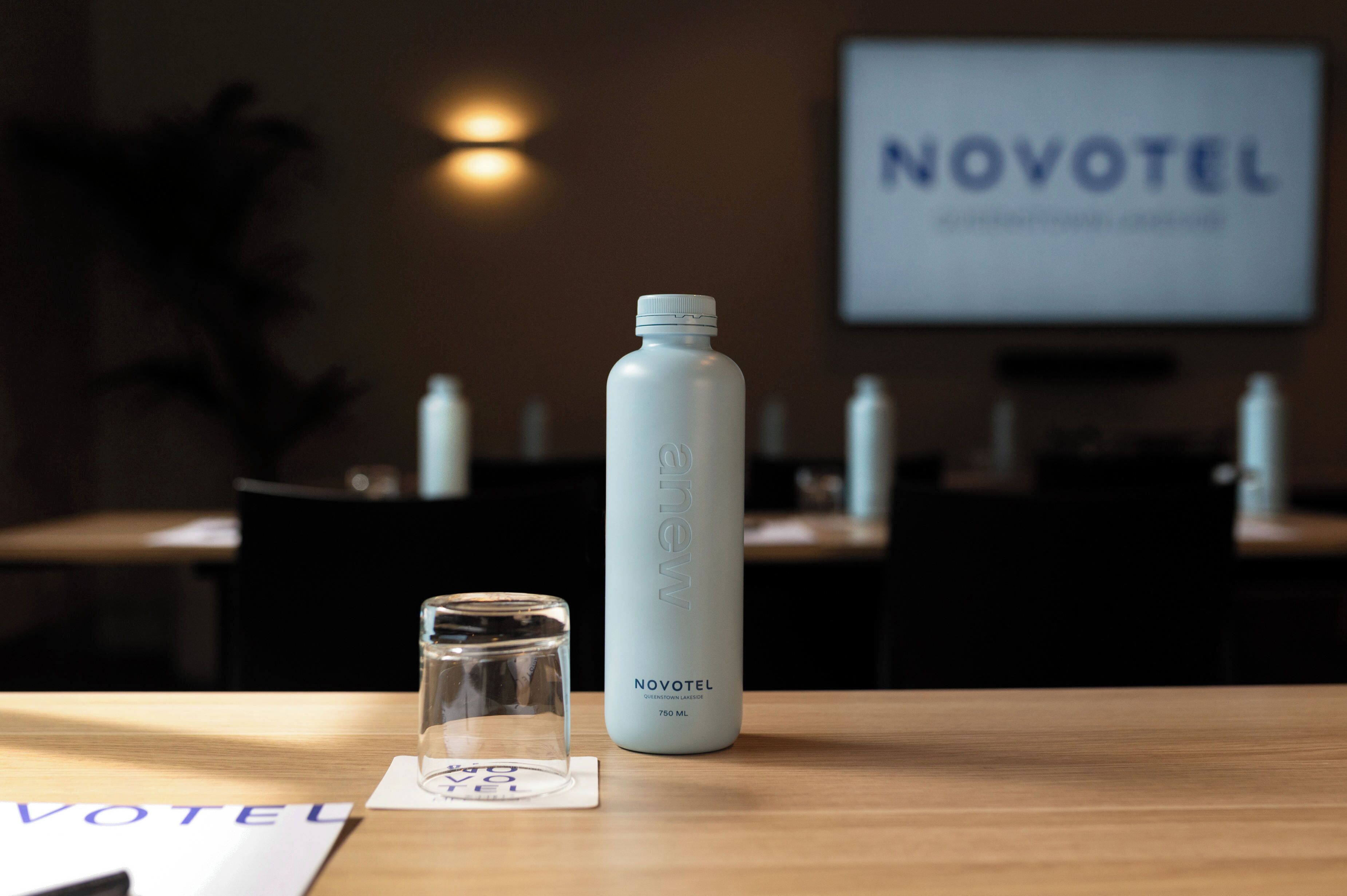 A hotel reception area with a water bottle and a glass on a wooden table, a sign reading "NOVOTEL", and a wall-mounted screen displaying the hotel name.