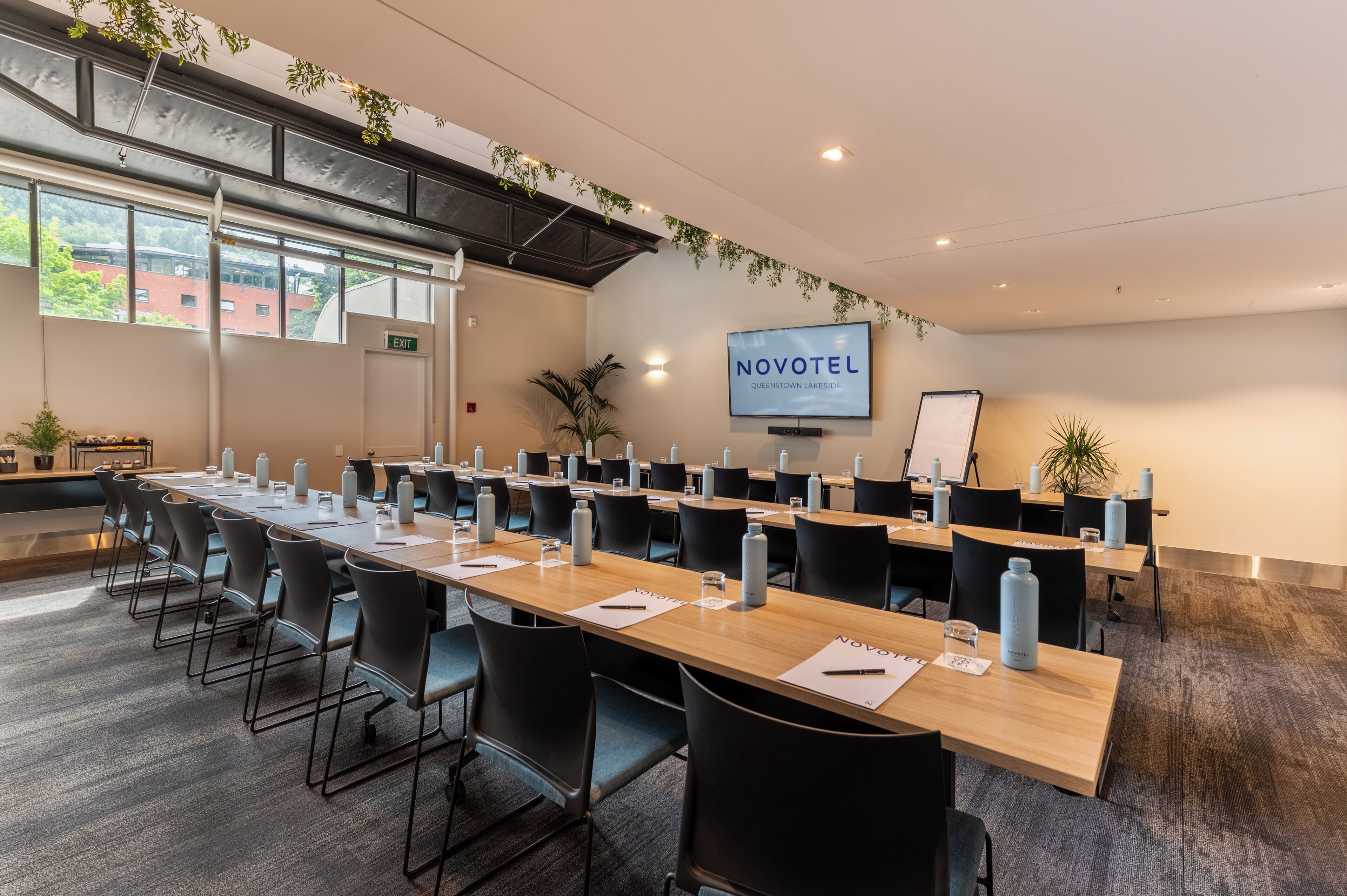 A conference room with long tables, black chairs, water bottles, and a TV screen displaying "NOVOTEL".
