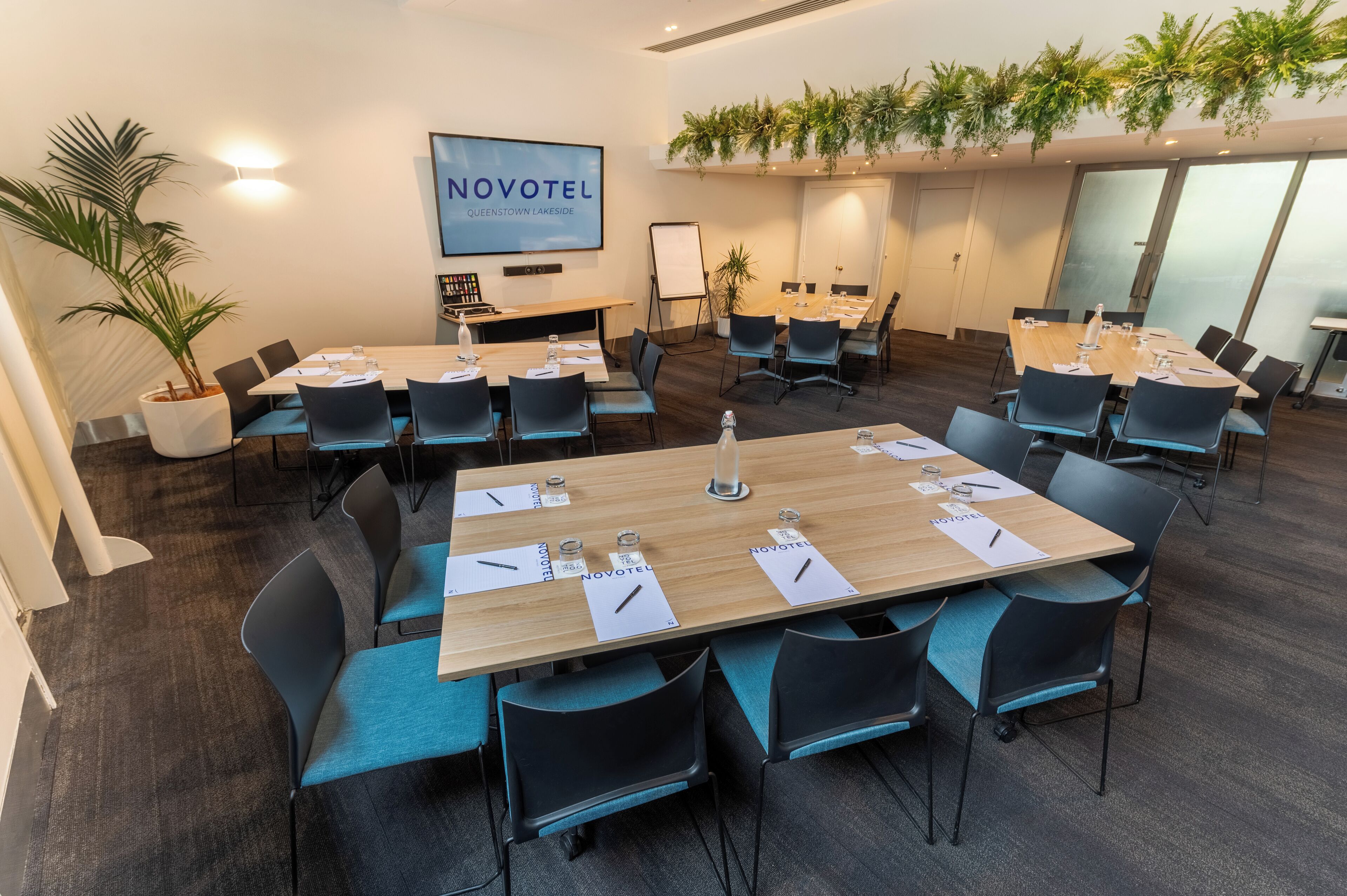 A conference room with multiple tables, chairs, and a large screen displaying "NOVOTEL QUEENSTOWN LAKESIDE".