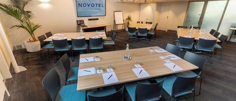 A conference room with multiple tables, chairs, and a large screen displaying "NOVOTEL QUEENSTOWN LAKESIDE".