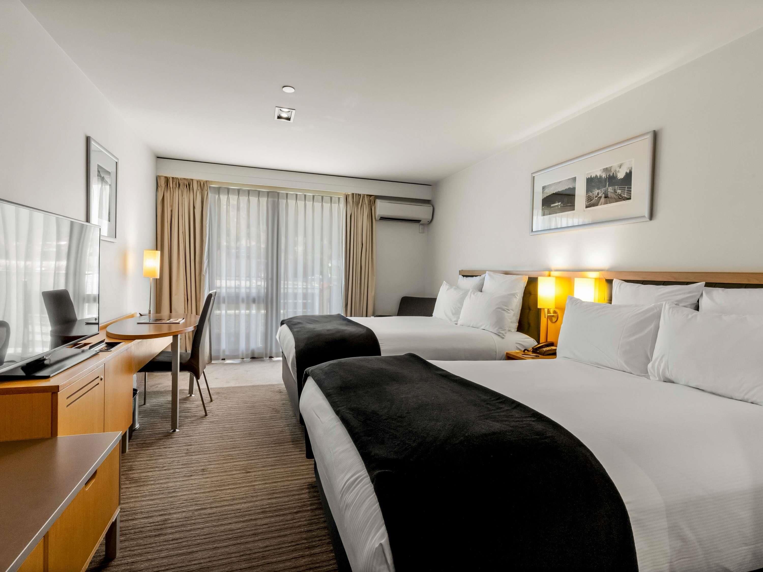 A hotel room with two beds, a desk, a chair, a TV, and a window with curtains.