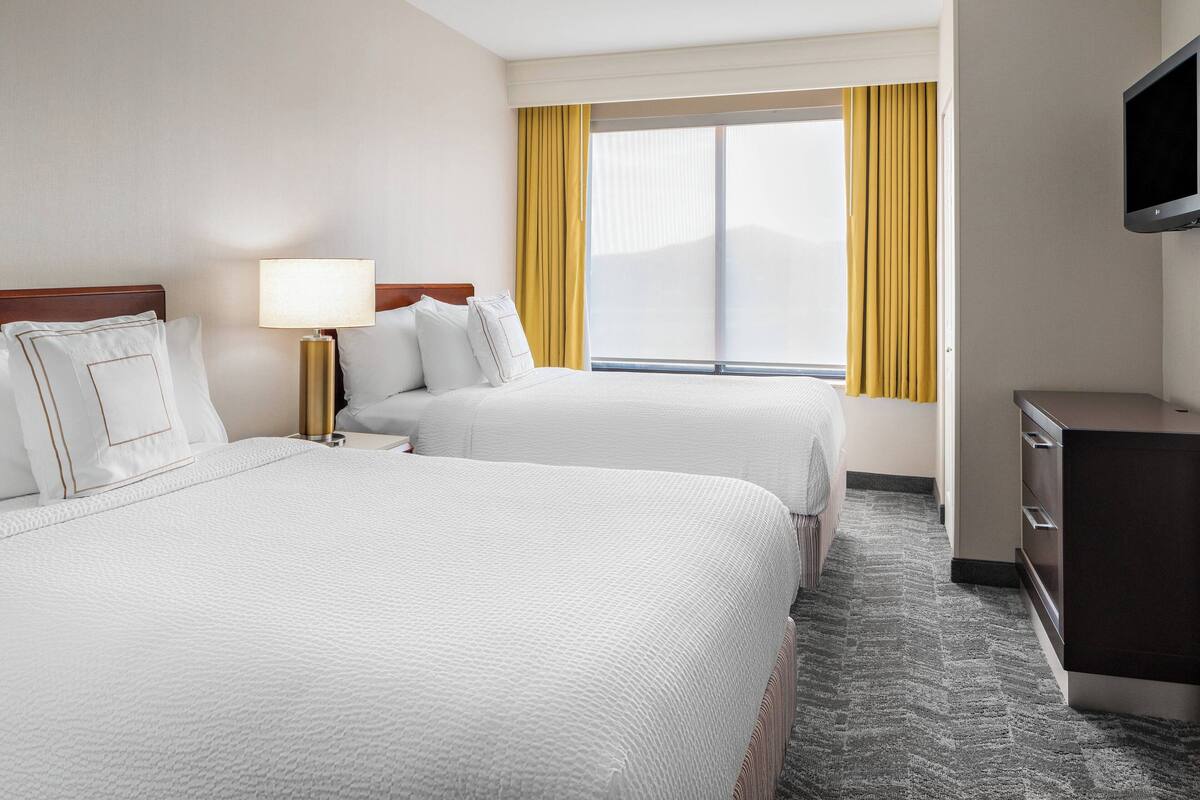 Ein Hotelzimmer mit zwei Betten, einem Fernseher, einem Schreibtisch und einem fenster mit Vorhängen.