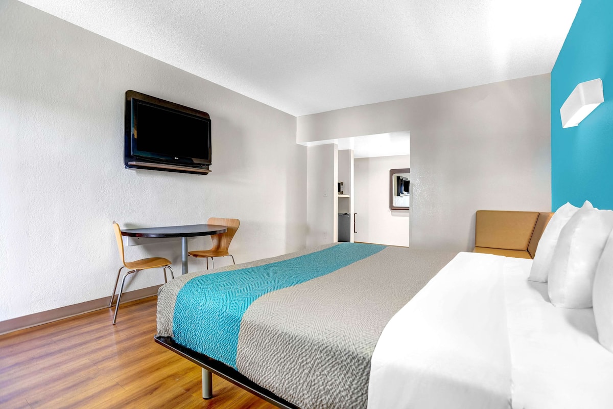 Habitación de hotel con cama, escritorio, silla, televisor y baño visible a través de una puerta abierta.