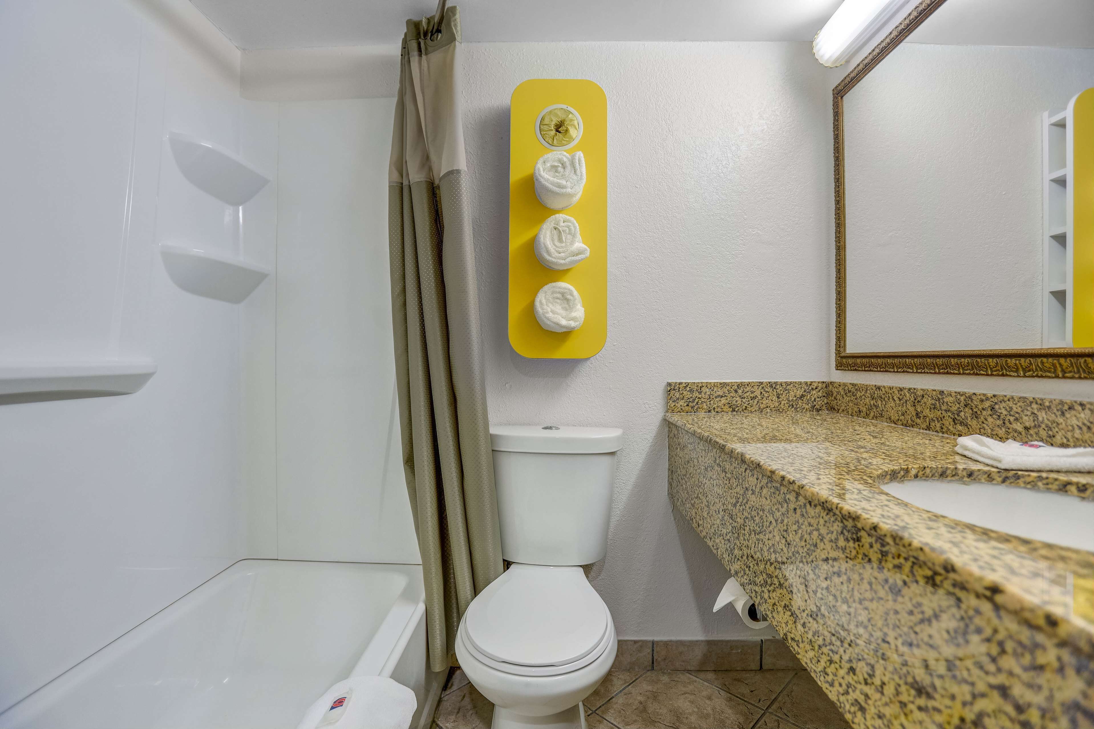 Un baño con un inodoro blanco, un toallero amarillo con cuatro toallas, una encimera de granito y un espejo.