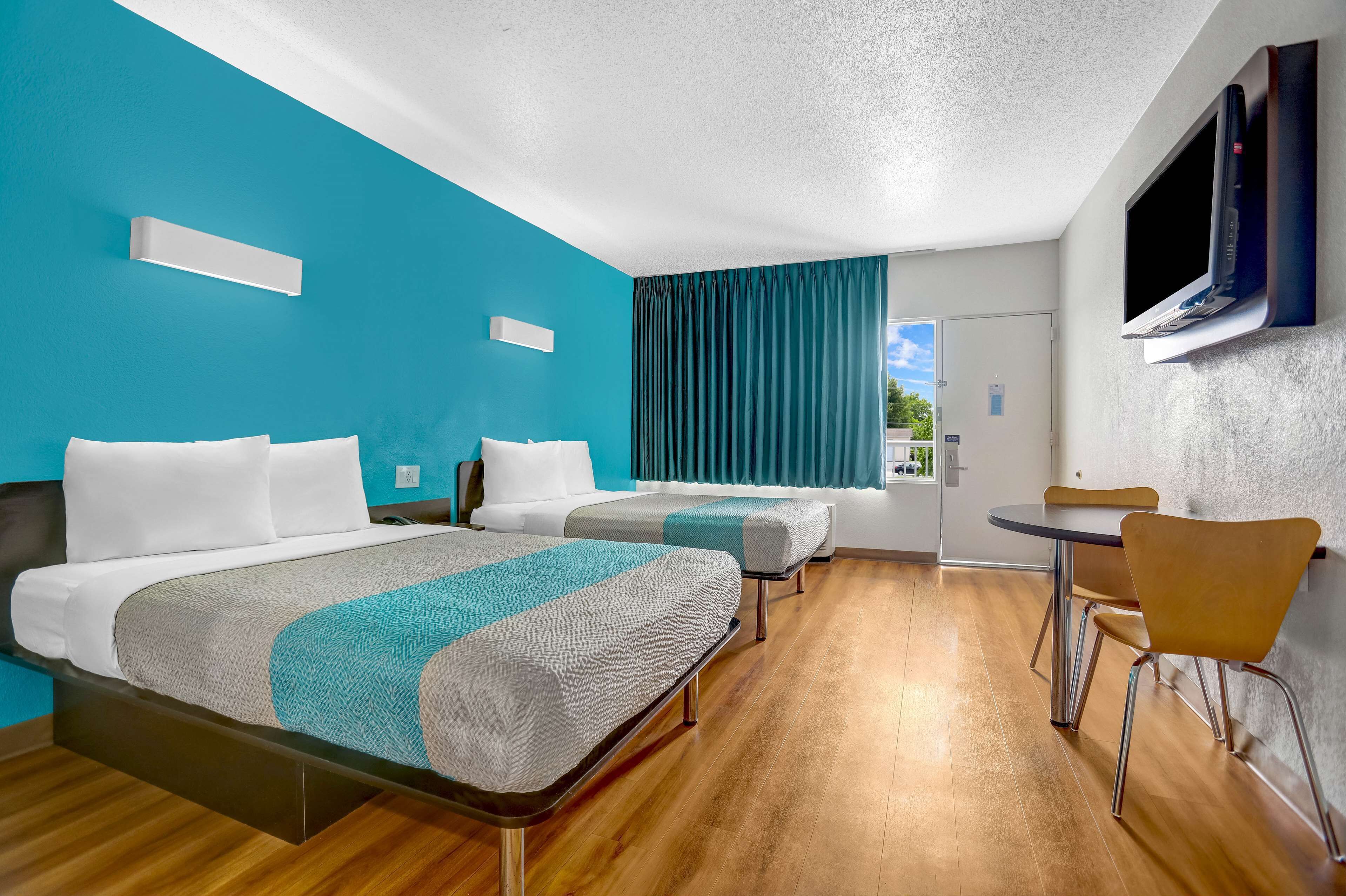 A hotel room with two beds, a small table, a chair, a TV, and a window with curtains.