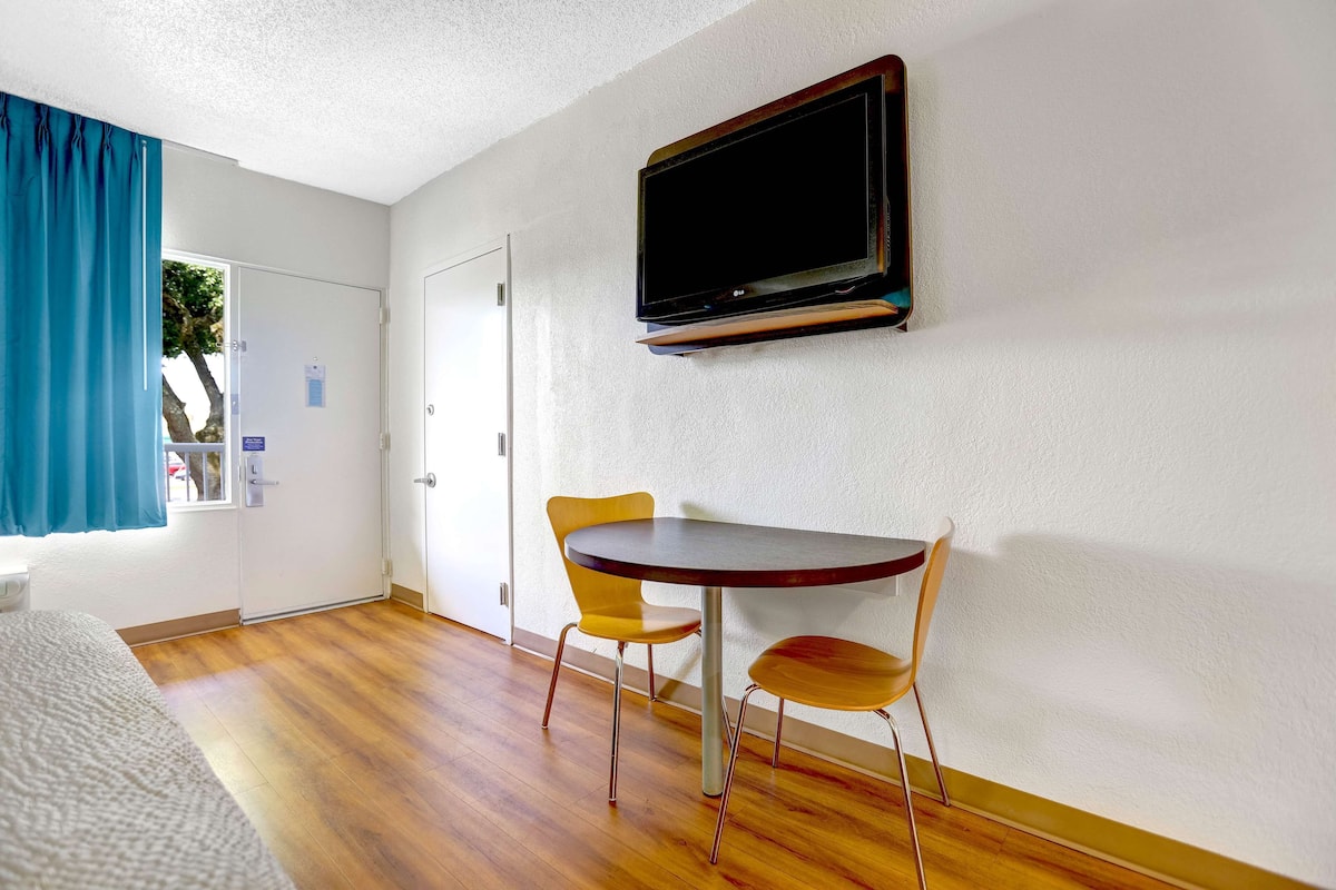 Una habitación de hotel con piso de madera, una cama, una mesa con dos sillas, un televisor montado en la pared y una ventana con cortinas azules.