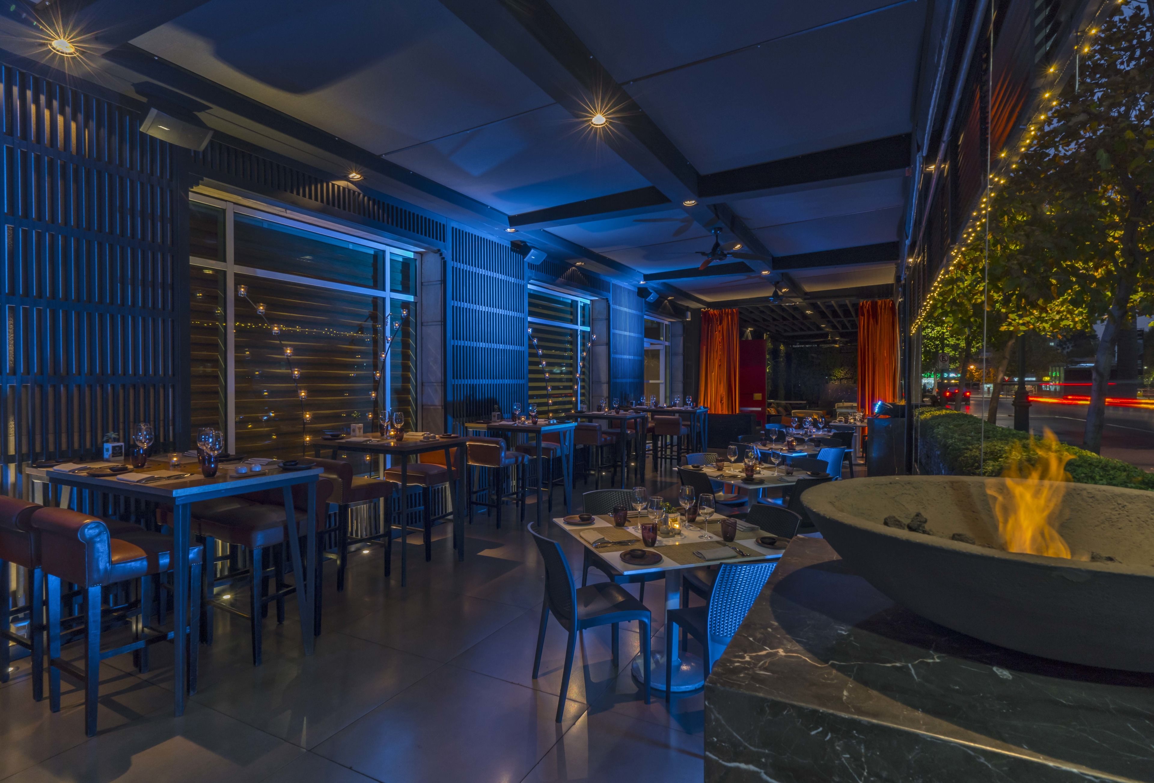 An indoor dining area with tables and chairs, a fire pit, and a view of the city at night.