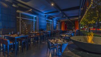 An indoor dining area with tables and chairs, a fire pit, and a view of the city at night.