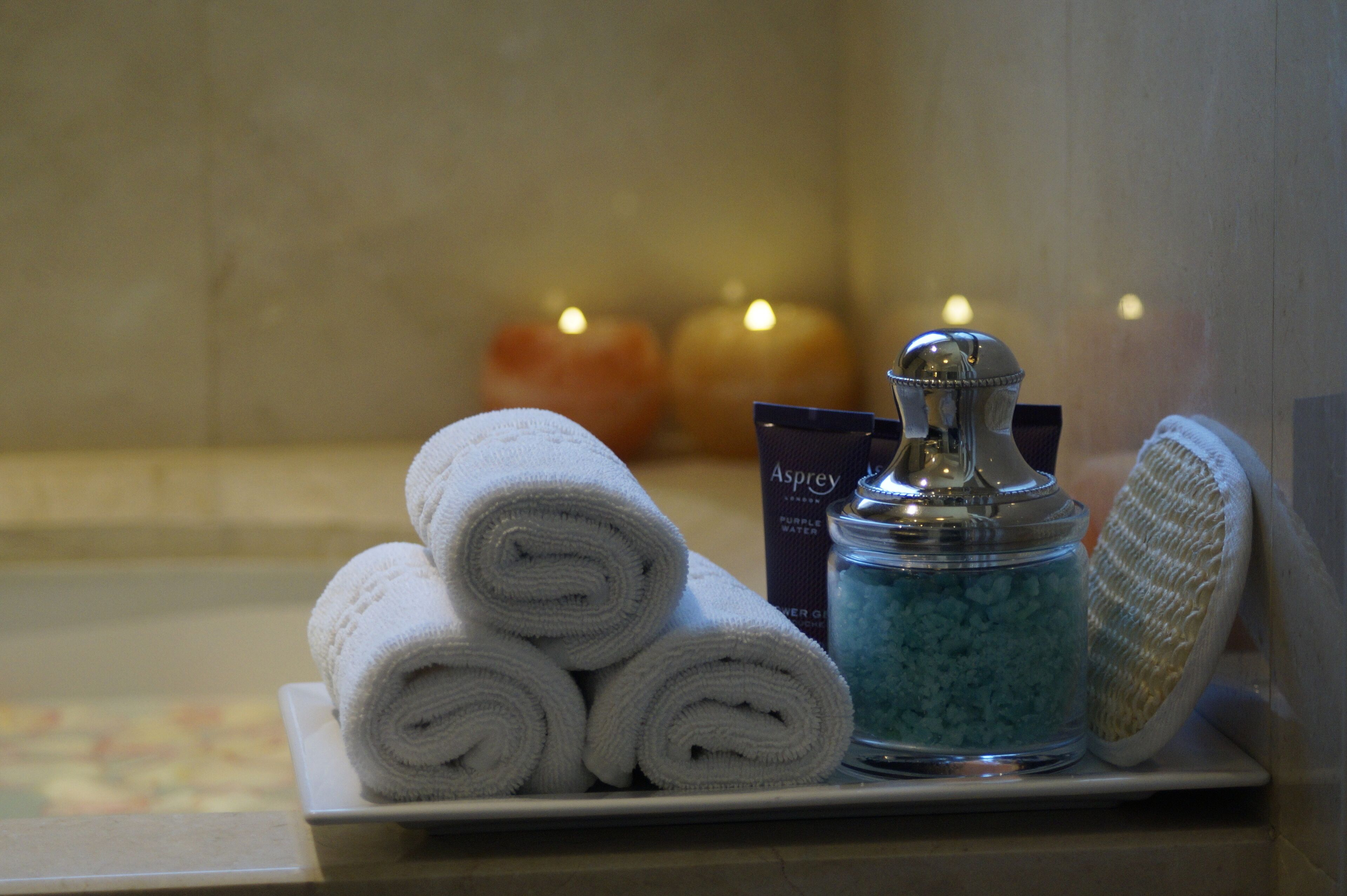 A bathroom with a towel rack holding rolled towels, a bottle of bath salts, and a bath brush.