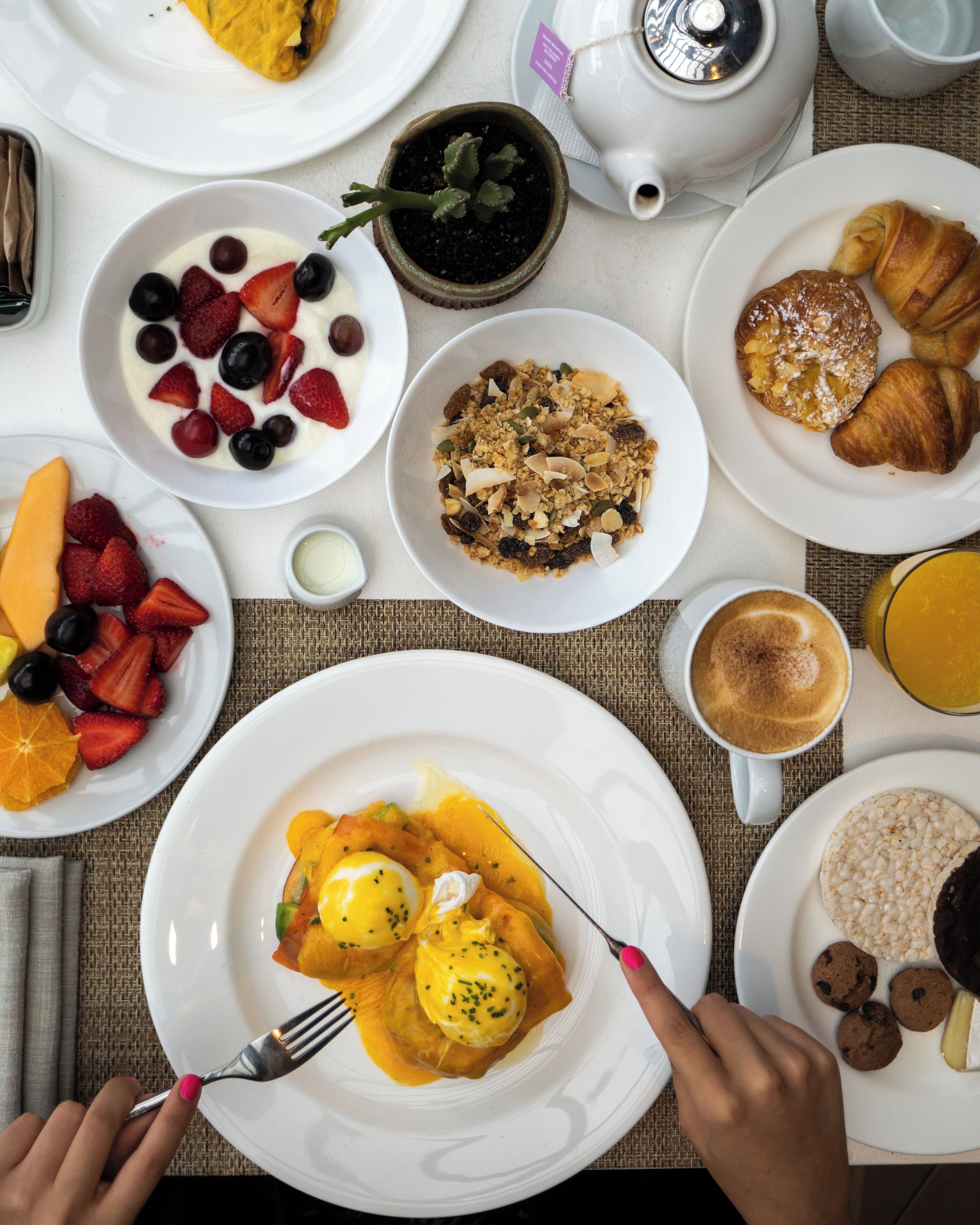 Una mesa de desayuno con diversos platos, entre ellos huevos benedictinos, croissants y fruta.