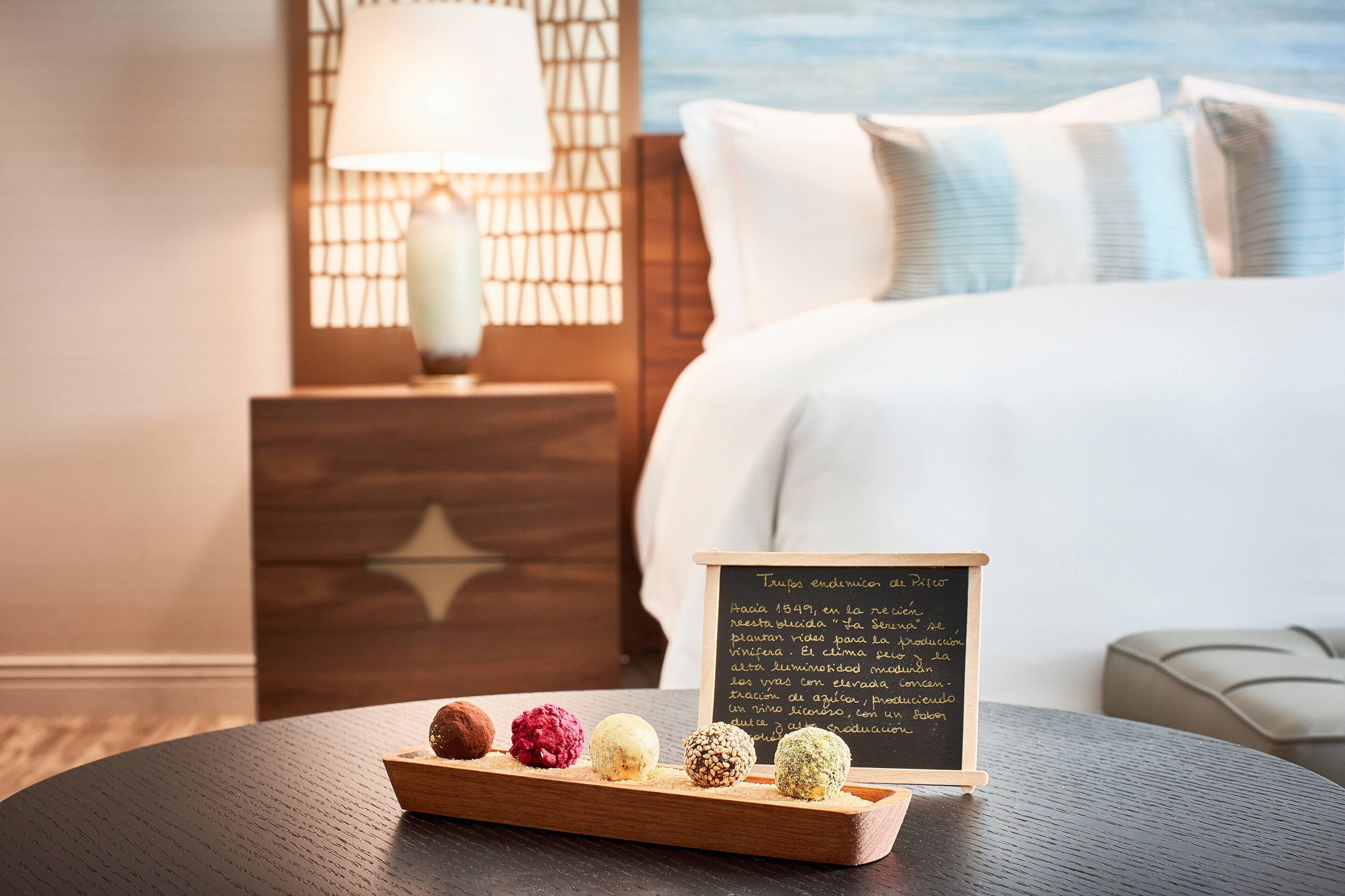 A neatly arranged tray of assorted chocolates on a round table with a wooden star design, a small chalkboard with text, and a bed with white bedding and blue pillows in the background.