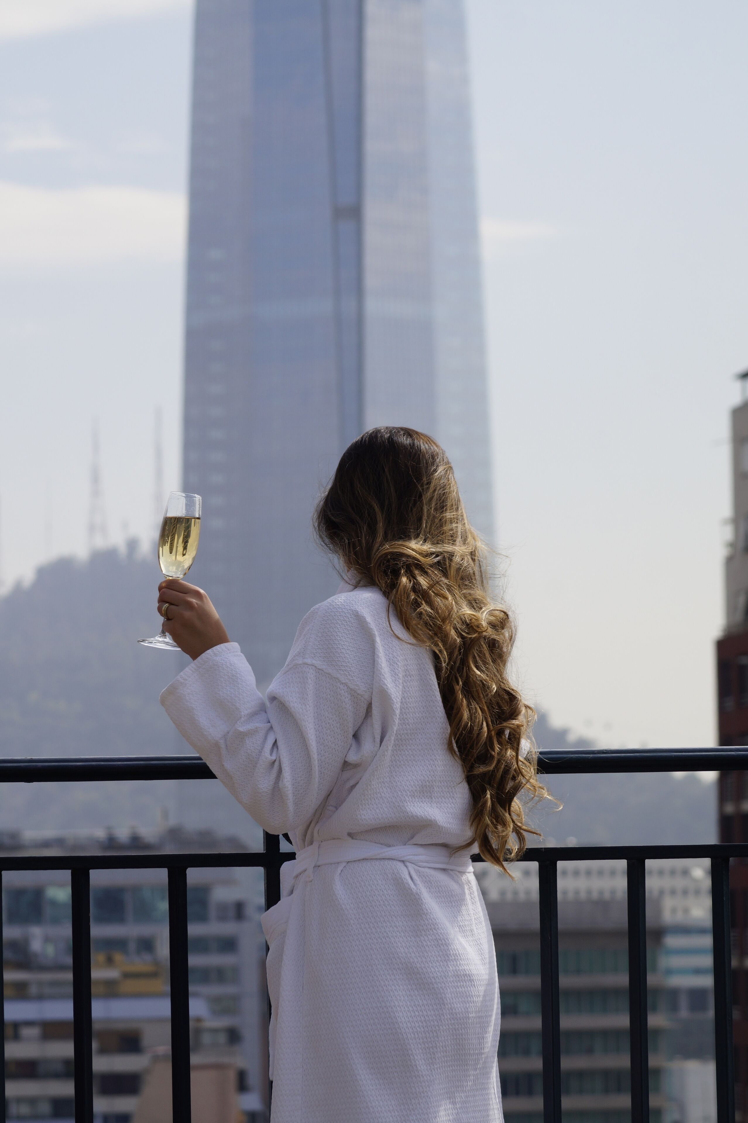 Una persona vestida con una bata blanca sostiene una copa de vino blanco, de pie en un balcón con vistas a un paisaje urbano y un edificio alto al fondo.