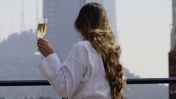 Una persona vestida con una bata blanca sostiene una copa de vino blanco, de pie en un balcón con vistas a un paisaje urbano y un edificio alto al fondo.
