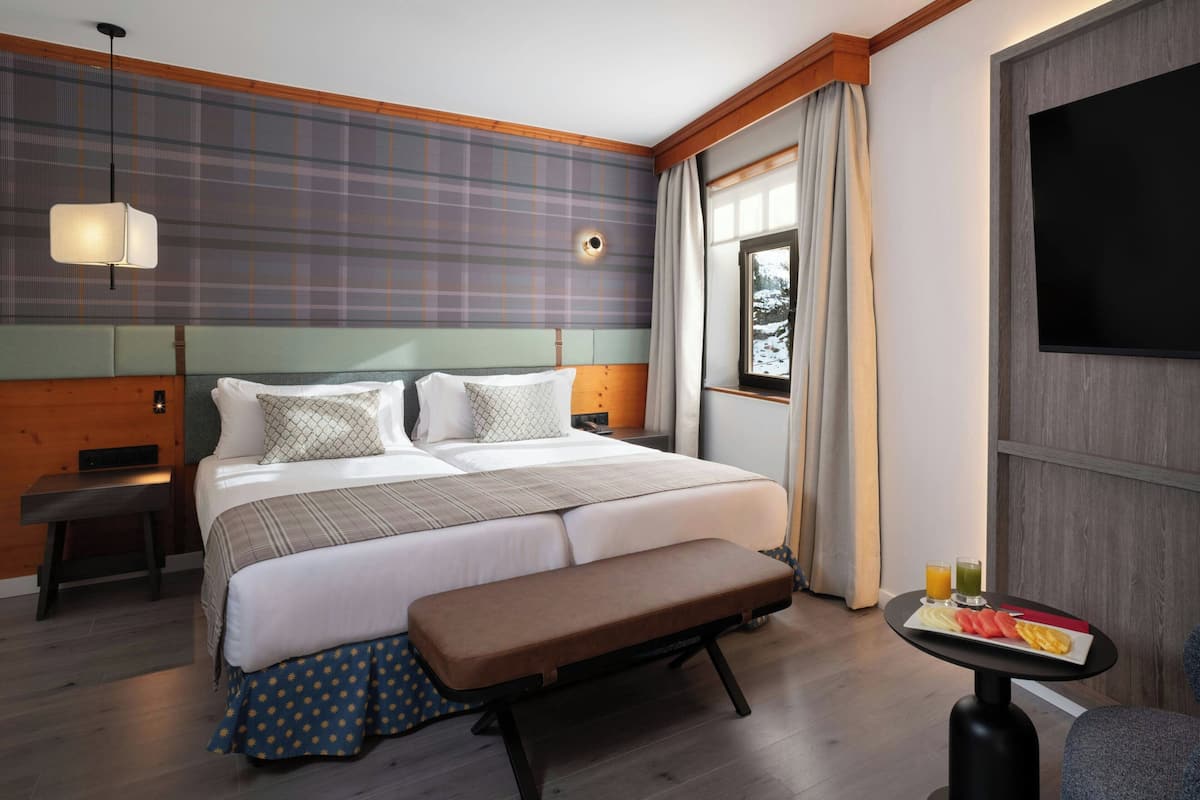 A hotel room with a large bed, a bench, a small table with breakfast, a TV, and a window with curtains.