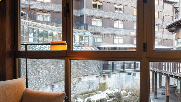 Une pièce chaleureuse avec un canapé, une petite table ronde sur laquelle se trouvent un livre et une tasse, et de grandes fenêtres donnant sur un bâtiment et un ruisseau.