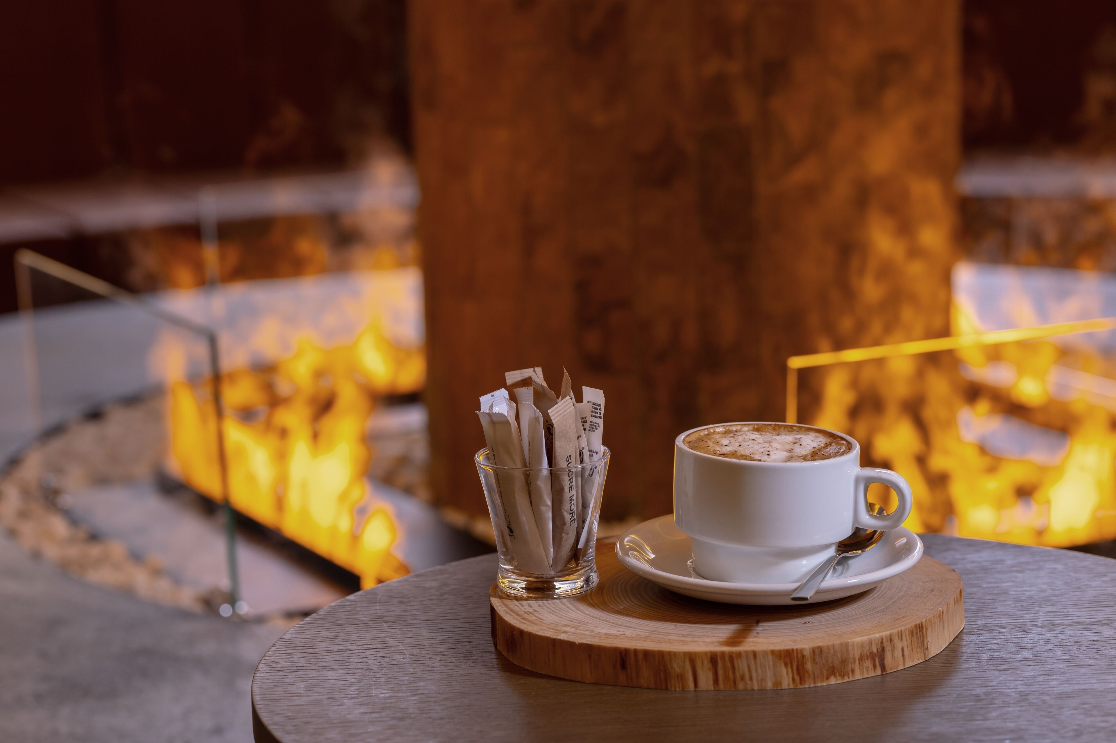 Una taza de café sobre un posavasos de madera, con un vaso de sobrecitos de azúcar a un lado, frente a un fondo con fuego.