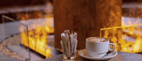 Una taza de café sobre un posavasos de madera, con un vaso de sobrecitos de azúcar a un lado, frente a un fondo con fuego.
