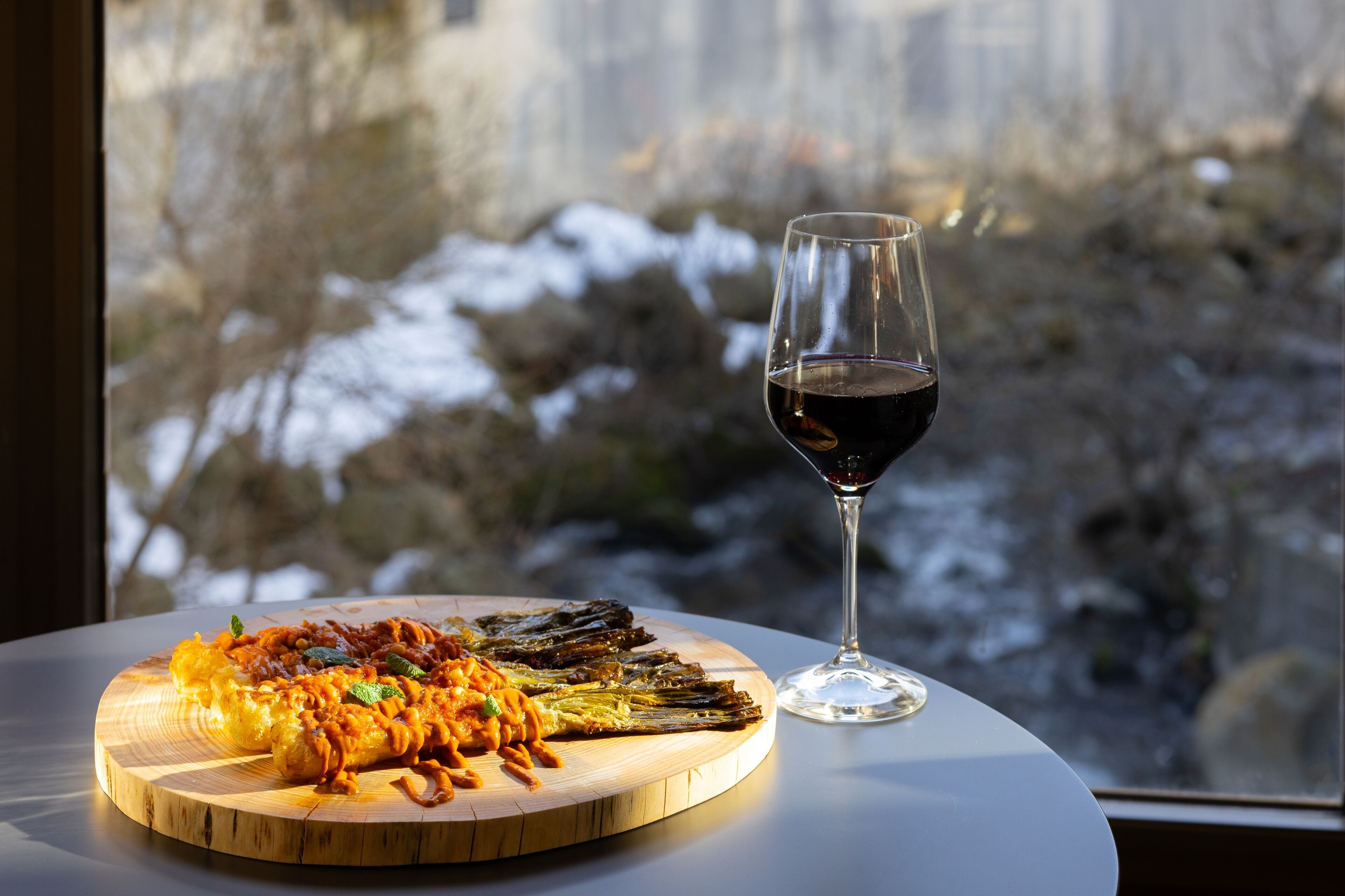 Una tabla de madera con pasta y verduras a la parrilla, una copa de vino tinto, y una vista de un paisaje nevado a través de una ventana.