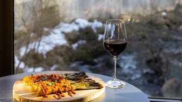 Una tabla de madera con pasta y verduras a la parrilla, una copa de vino tinto, y una vista de un paisaje nevado a través de una ventana.