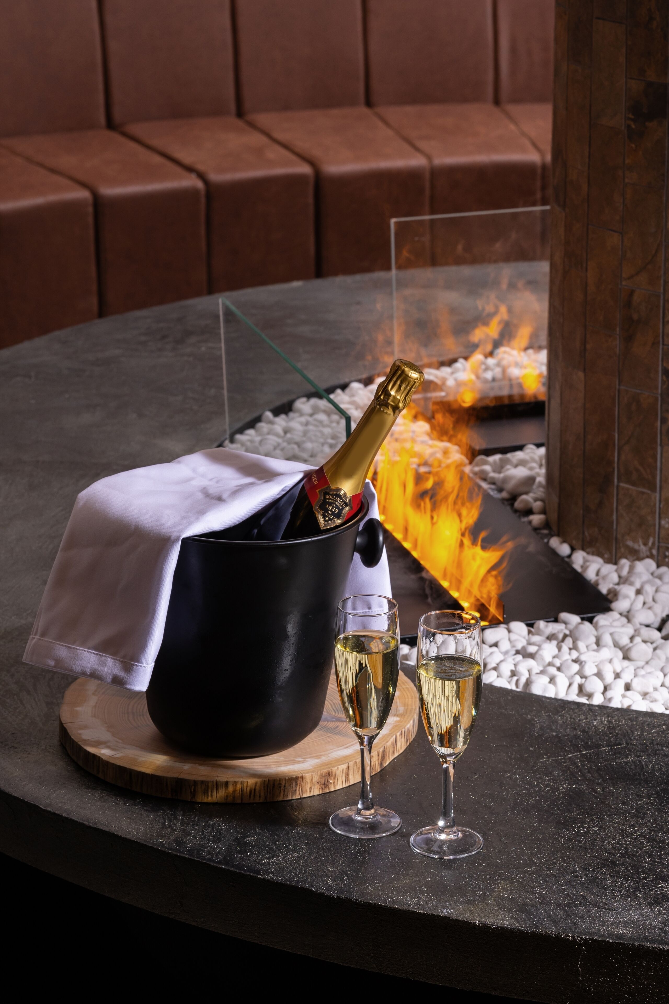 Una botella de champán en un cubo negro con tela blanca, dos copas de champán y una chimenea decorativa al fondo.
