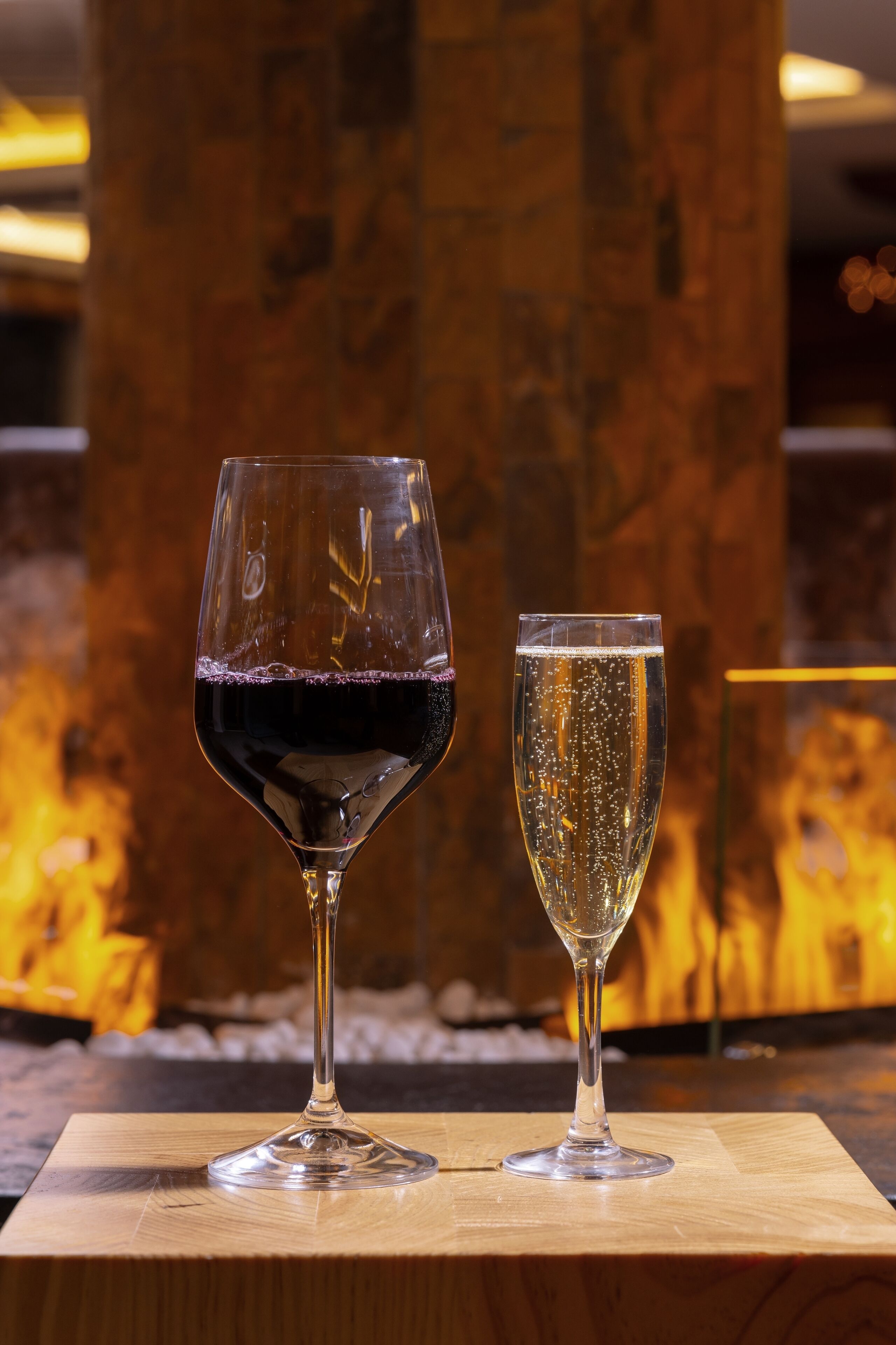Deux verres à vin sont posés sur une table en bois, devant un mur de pierres et avec une cheminée allumée en arrière-plan.