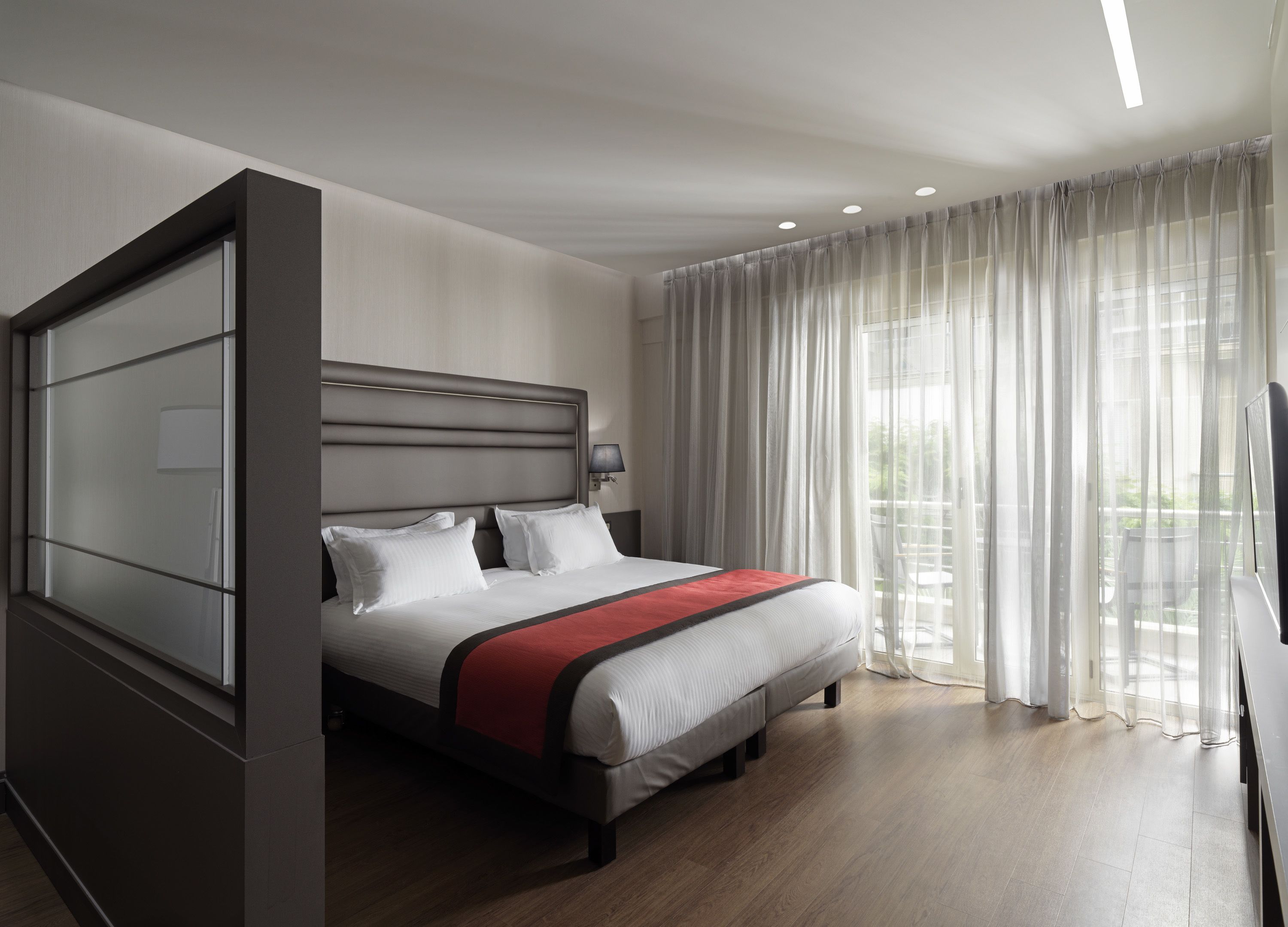 A modern hotel room with a large bed, a bedside table, a lamp, and sheer curtains.
