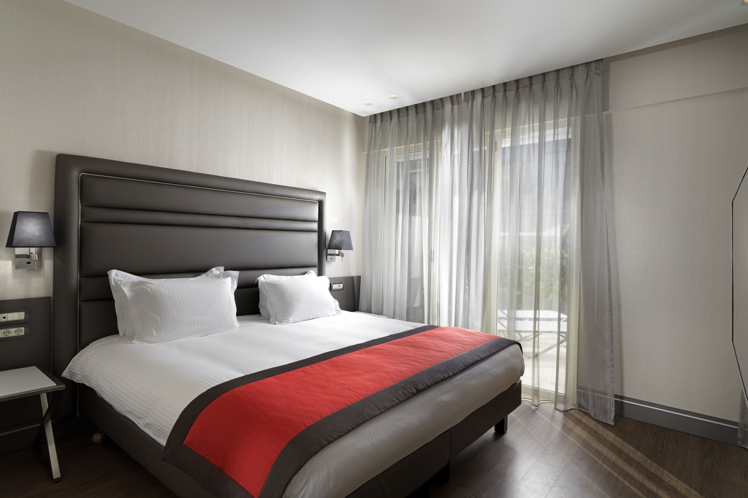 A modern hotel room with a large bed, a nightstand, a lamp, and a view of the outdoors through sheer curtains.
