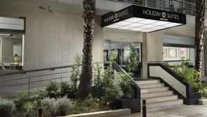 L’entrée d’un hôtel, avec une enseigne indiquant « Holiday Suites » et « Arnis Street ». Des marches mènent à l’entrée, qui est entourée de plantes et d’une rambarde.