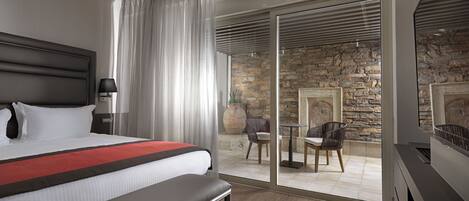 A modern hotel room with a large bed, a bench, a desk, and a stone wall.