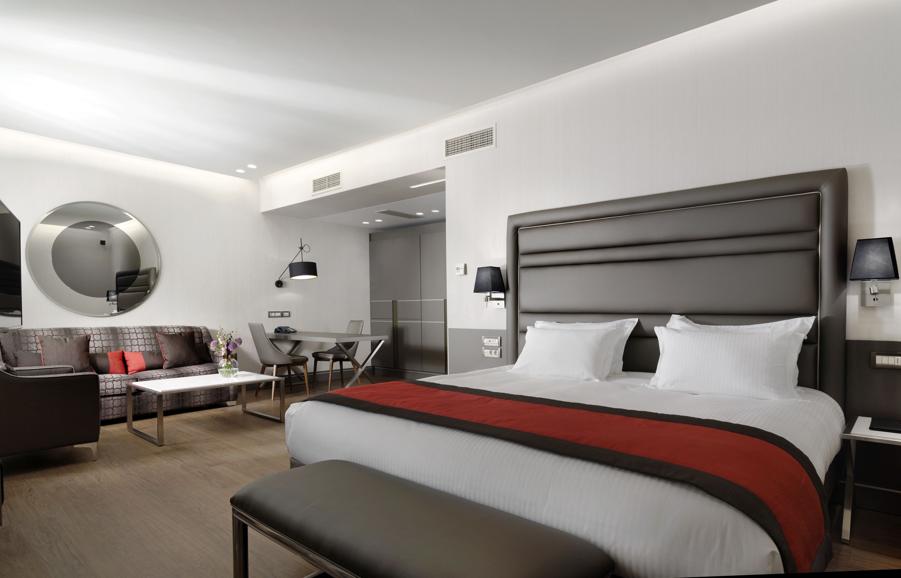 A modern hotel room with a large bed, a sofa, a desk, and a mirror.