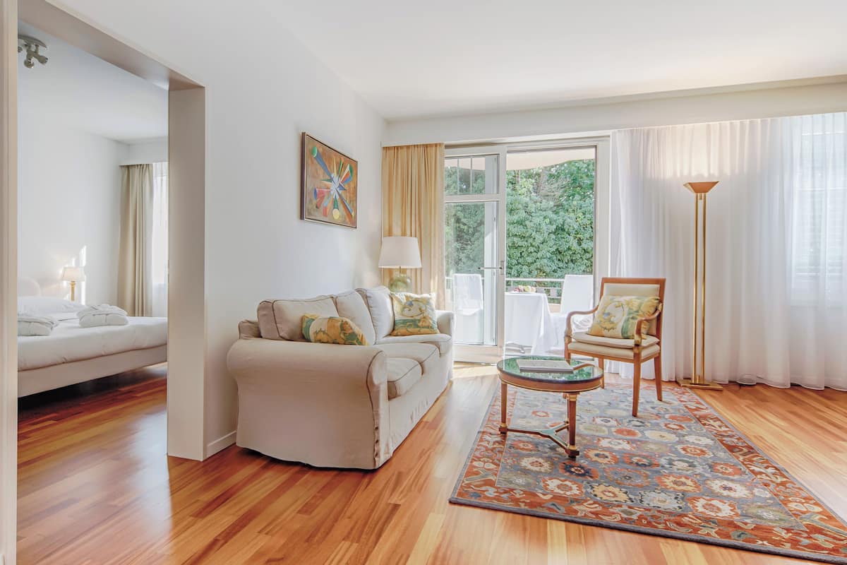 A spacious living room with a sofa, armchair, and a coffee table, a large window with curtains, and a painting on the wall.