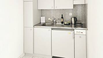 Cocina compacta con armarios blancos, pileta de acero inoxidable, tetera y lavavajillas.