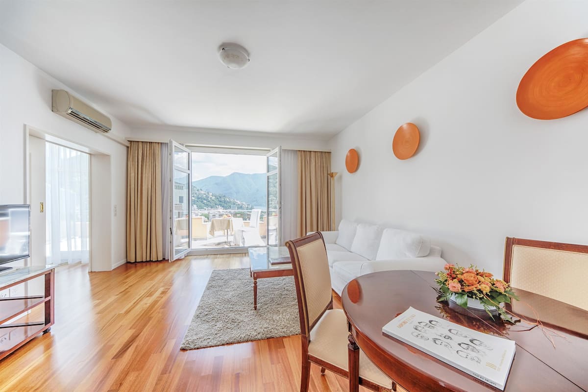 A modern hotel room with a wooden floor, a white sofa, a dining table with a vase of flowers, and a large window offering a city view.