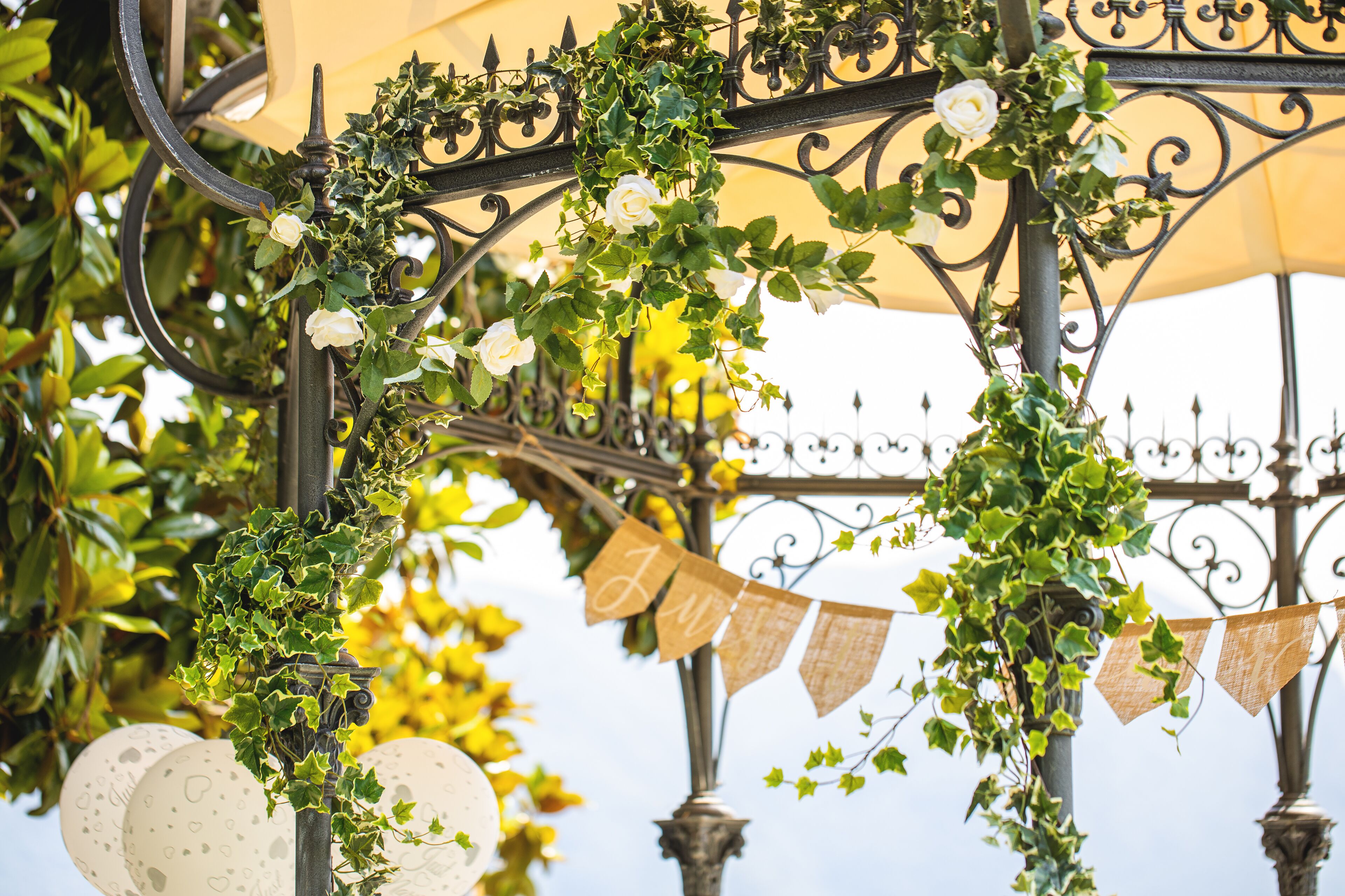 A wrought iron structure adorned with ivy and white roses, with bunting flags and decorative plates.