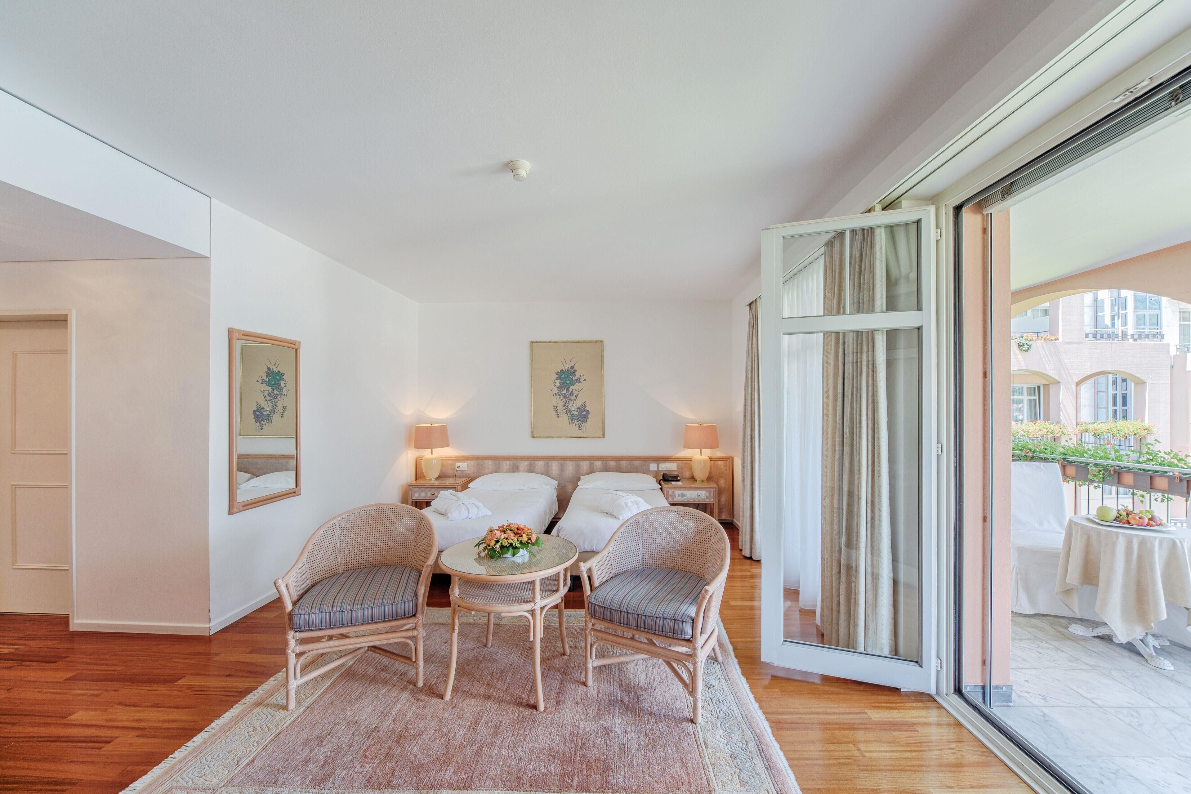 Ein Hotelzimmer mit einem Bett, zwei Stühlen, einem kleinen Tisch mit Blumen und einem Balkon mit Tisch und Stühlen.