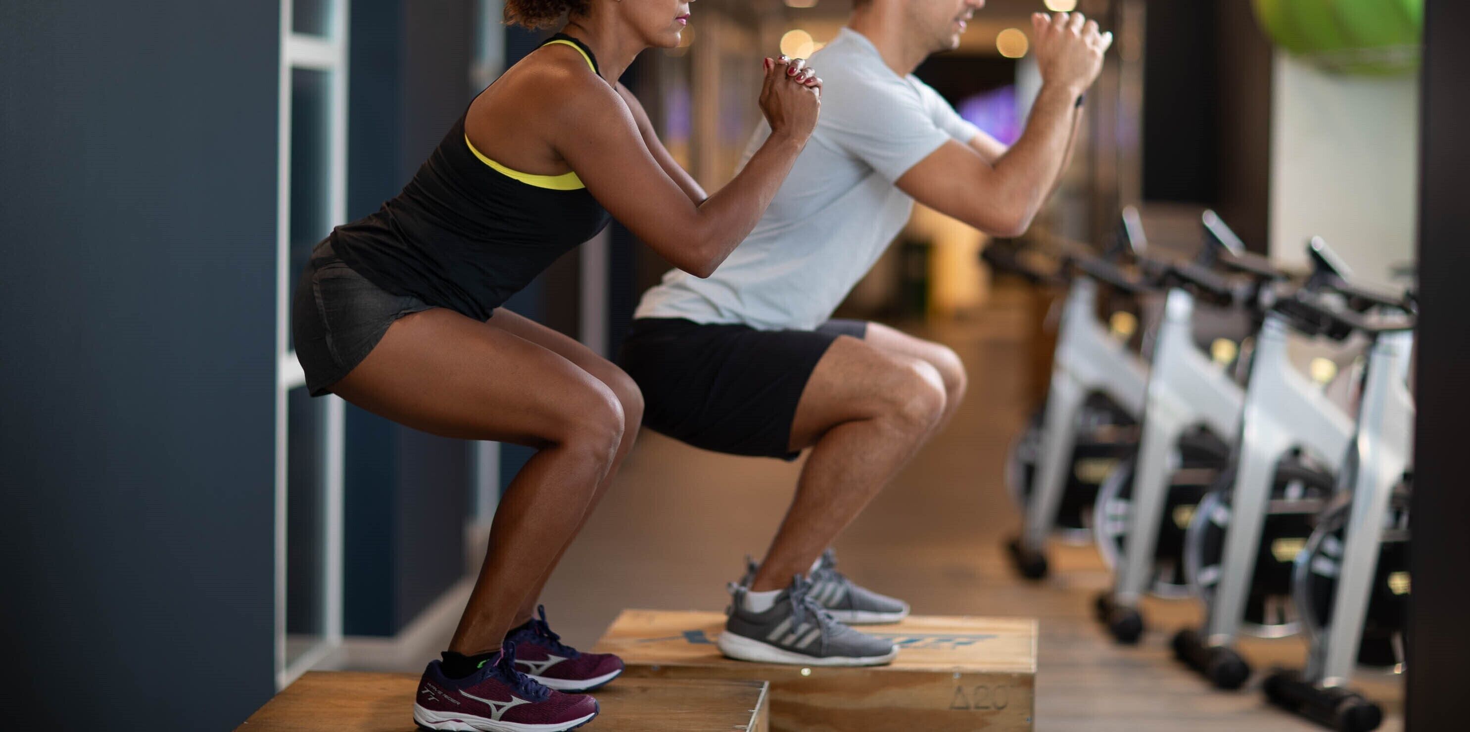 Two people performing squats in a gym with exercise equipment in the background.