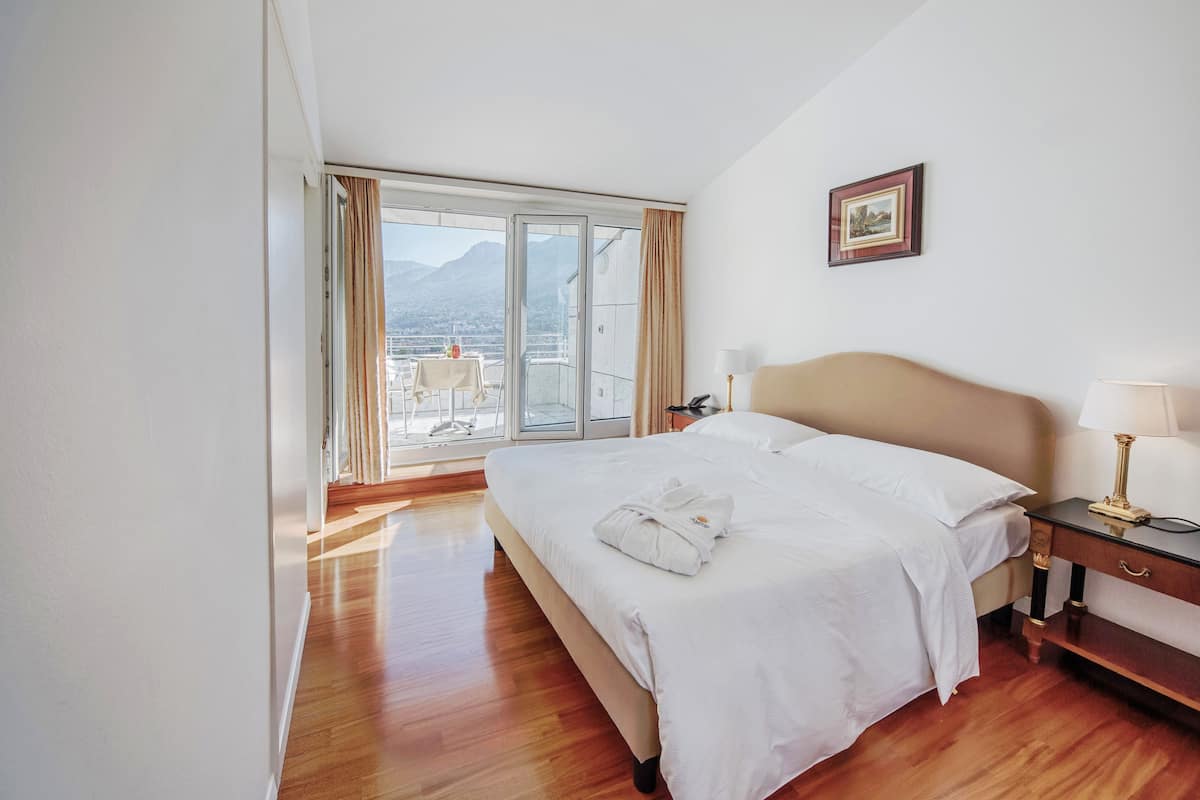 Habitación de hotel con una cama grande, una mesita de noche con lámpara, vista a montañas a través de una puerta corrediza de vidrio y un cuadro en la pared.