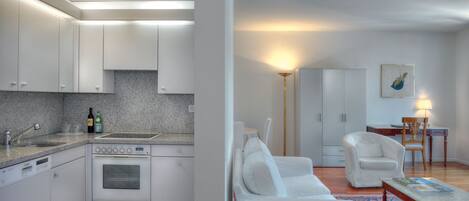 Una cocina moderna con armarios blancos, horno integrado y encimera de granito. Hay una zona de comedor con sofá blanco y alfombra estampada.