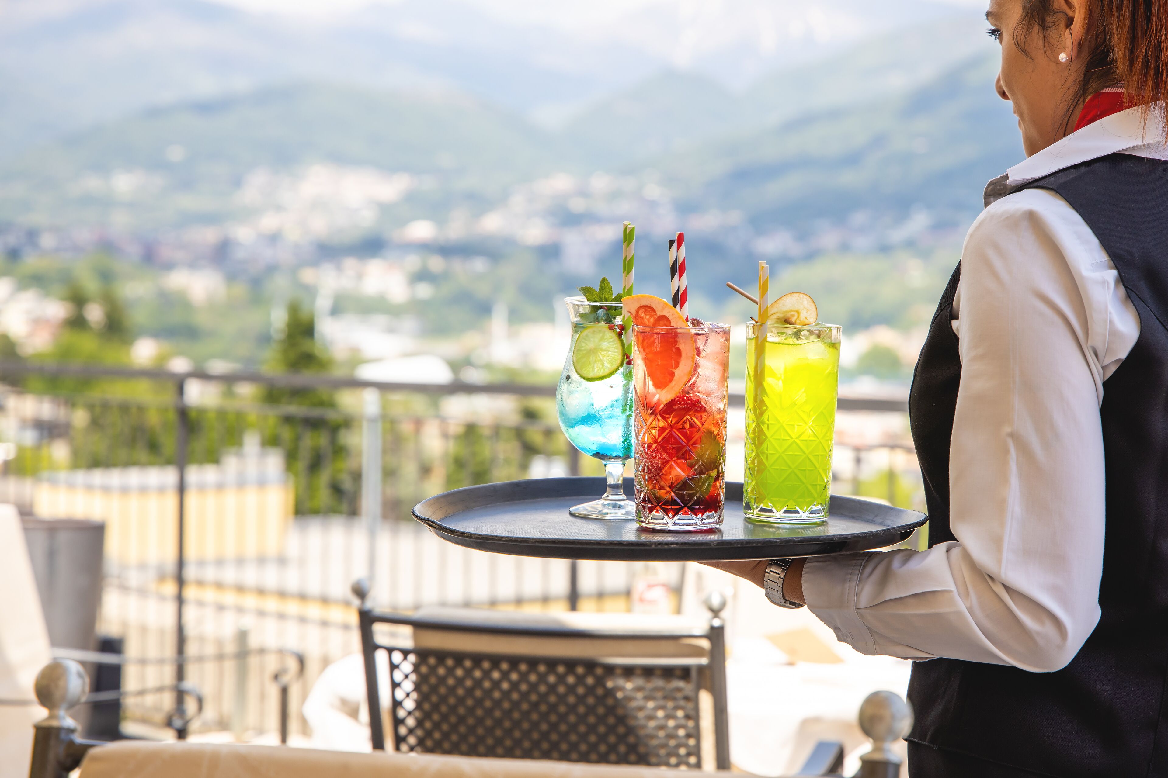 A person holding a tray with three colorful drinks, a cityscape with mountains in the background, and outdoor seating.