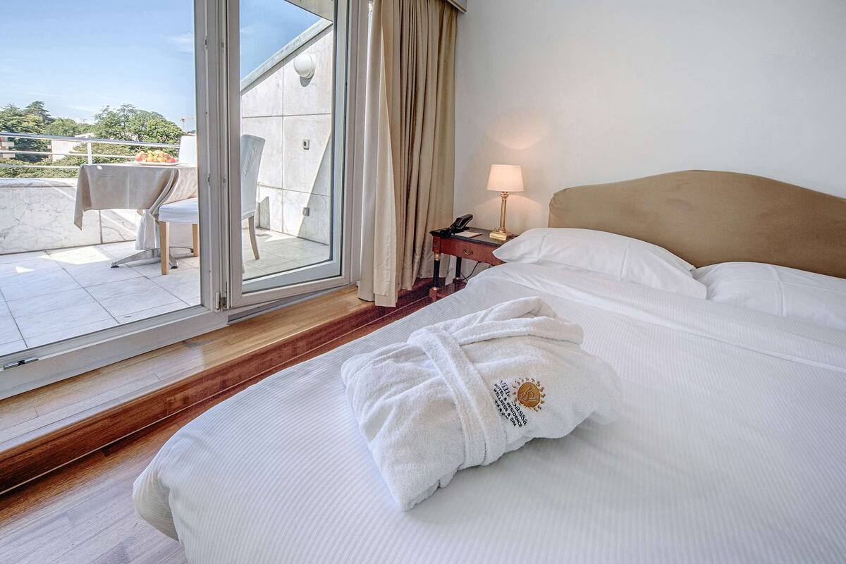 A hotel room with a bed, a nightstand, a lamp, a robe, and a sliding glass door leading to an outdoor seating area.