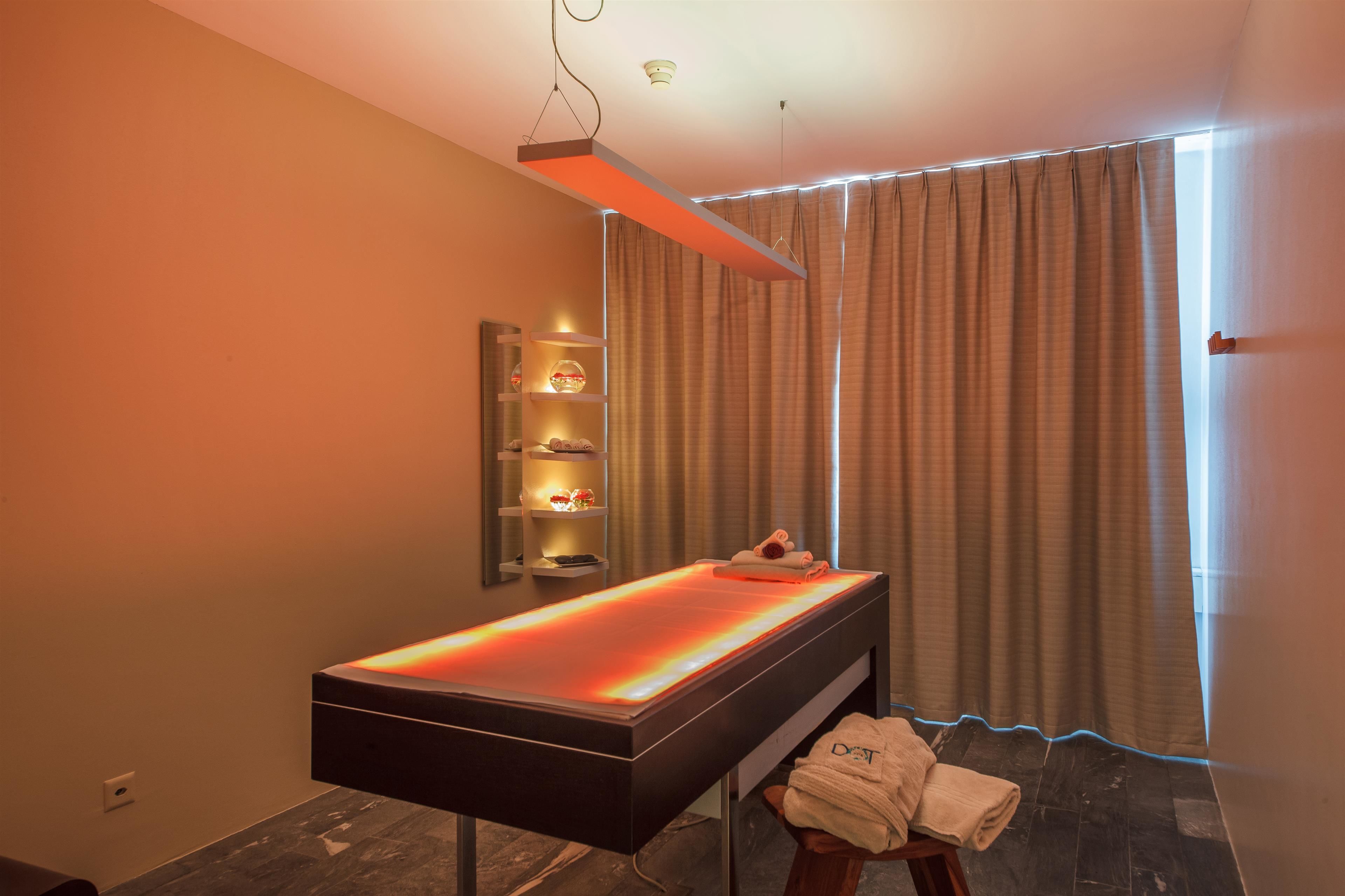 Une salle de spa moderne équipée d’une table de massage chauffante, d’une étagère avec des bougies et offrant une vue à travers des rideaux.