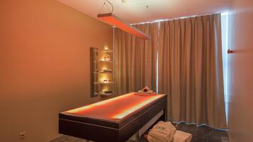 Ein moderner Spa-Raum mit einer beheizten Massageliege, einem Ablagebereich mit Kerzen und Blick durch Vorhänge.