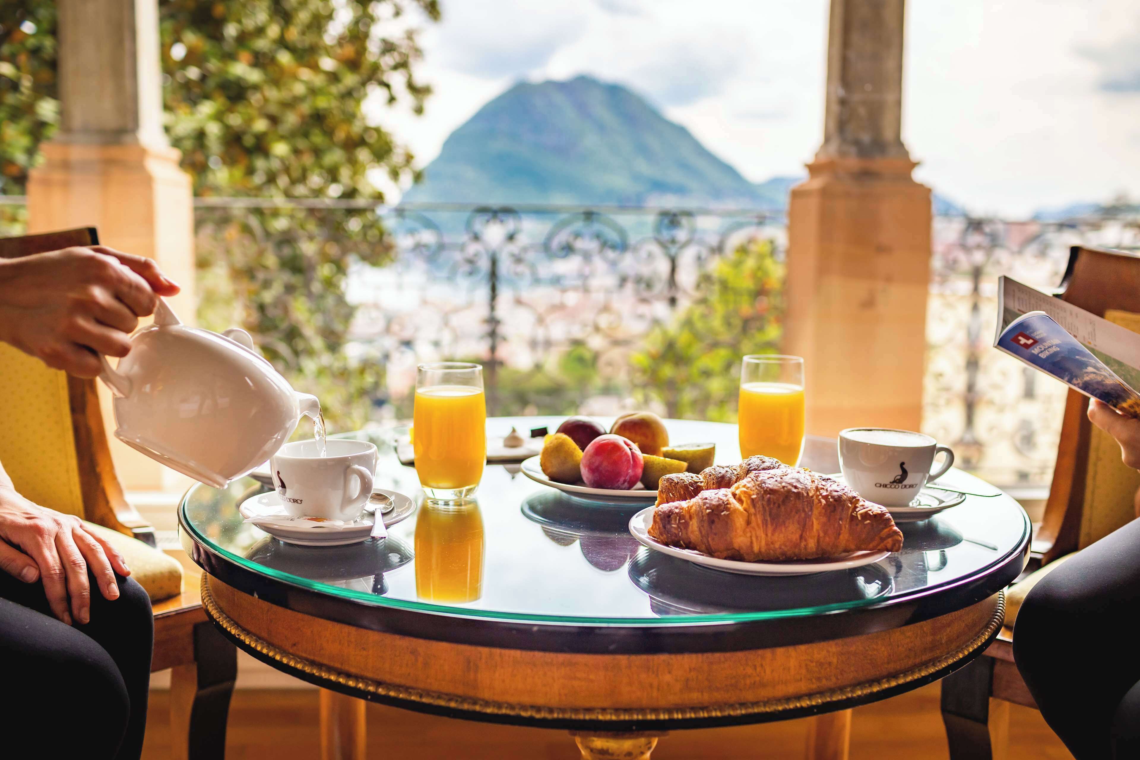 Una mesa de desayuno con un croissant, fruta, jugo y café, con vistas a un paisaje escénico que incluye una montaña al fondo.
