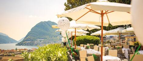 An outdoor dining area with tables and chairs, umbrellas, and a scenic view of a lake and mountains.
