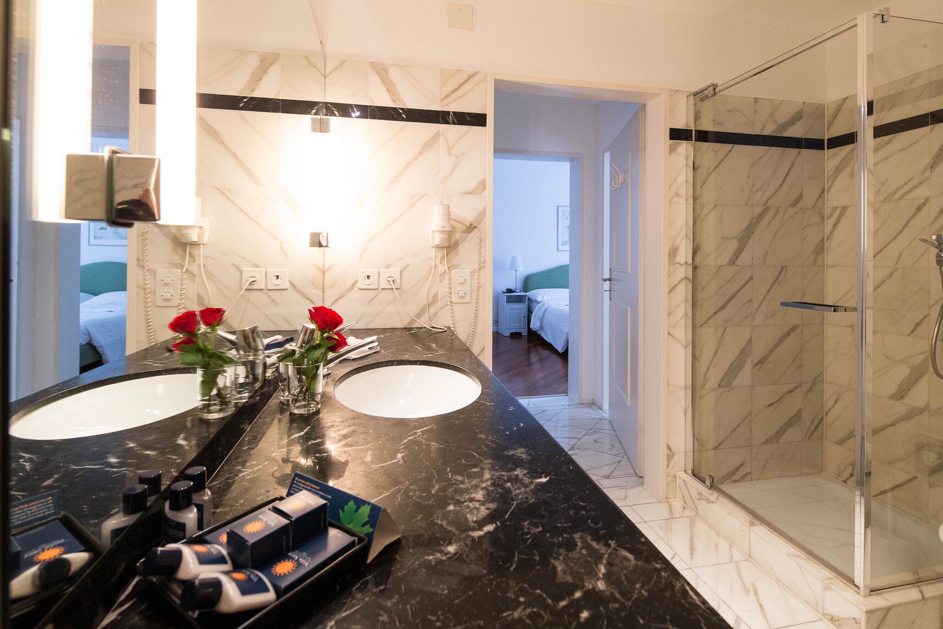 Un bagno con due lavandini, uno specchio, cabina doccia in vetro e un vaso con fiori rossi sul mobile.