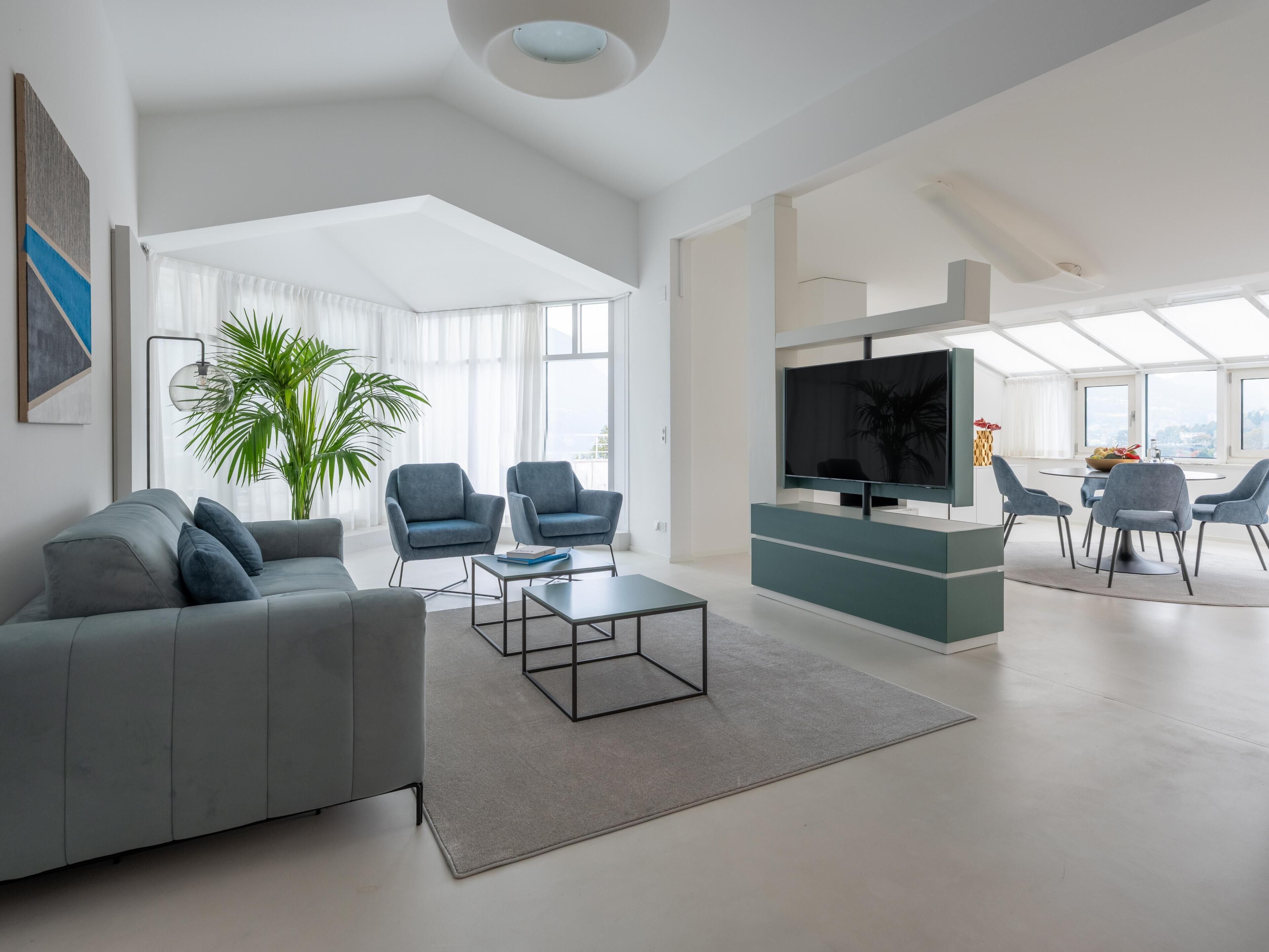 Una sala de estar moderna con un sofá gris, dos sillones, una mesa de centro, un televisor y una zona de comedor con sillas y una mesa.
