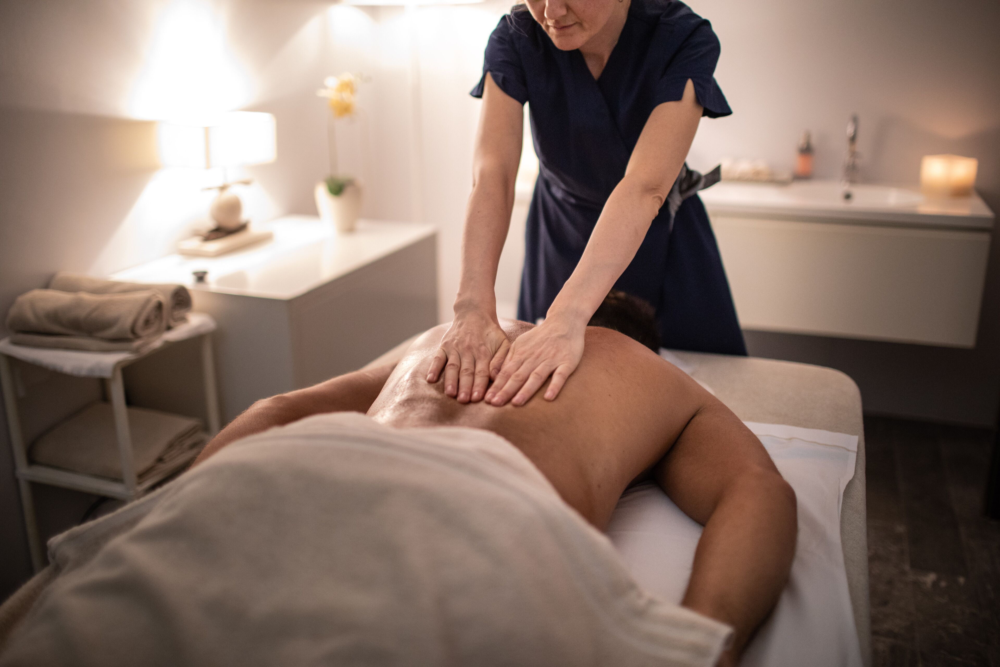 Sesión de masajes en un spa, con un cliente acostado en una camilla de tratamiento, un terapeuta atendiendo y una zona de recepción con mostrador y lámpara al fondo.