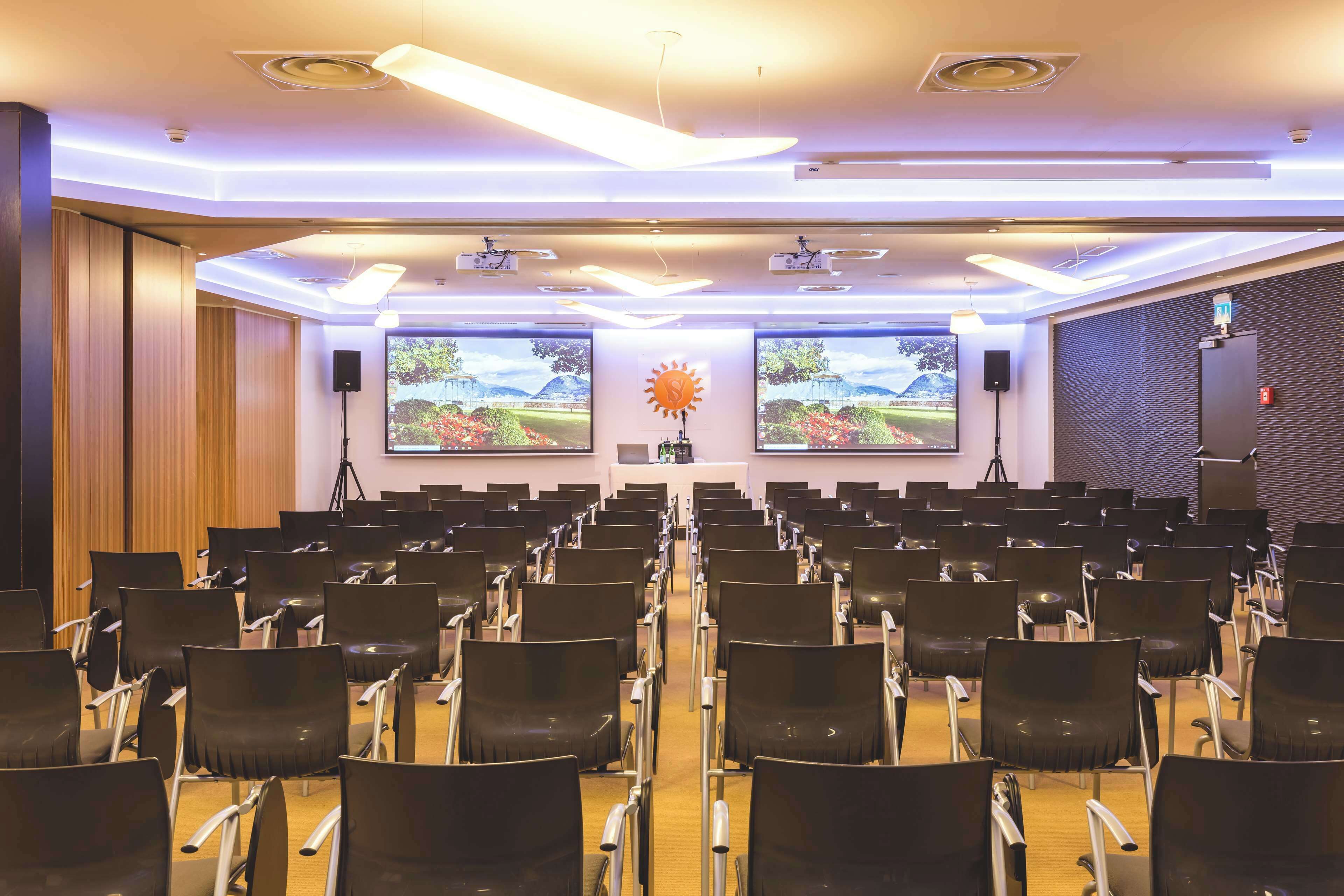 Sala de conferências com fileiras de cadeiras, um púlpito, duas telas grandes e uma decoração em formato de sol.