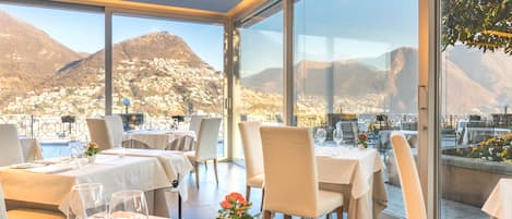 A restaurant with tables set for dining, large windows offering a view of mountains and a town, and a modern interior design.