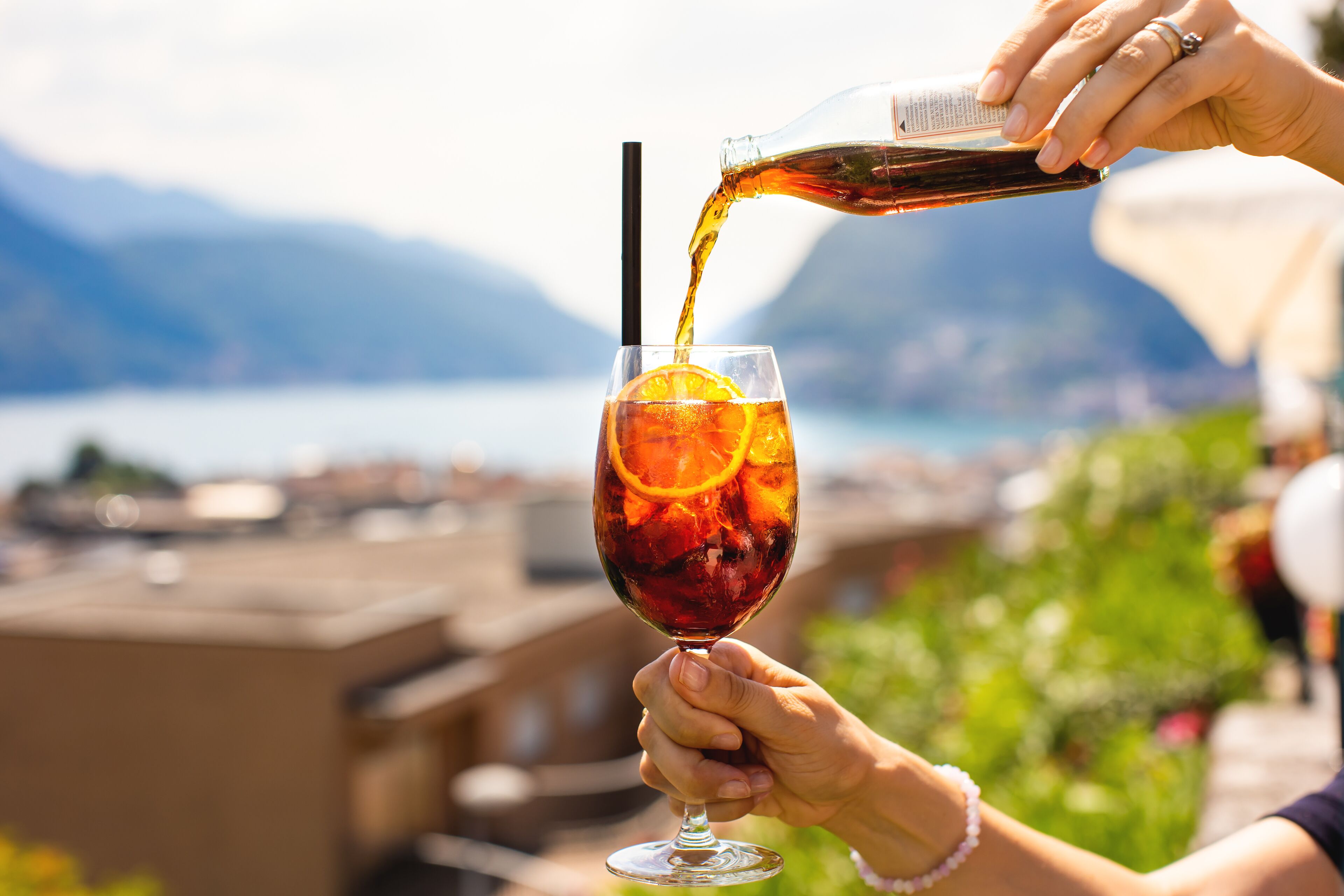 A person pouring a drink into a glass with an orange slice, with a scenic view of mountains and water in the background.
